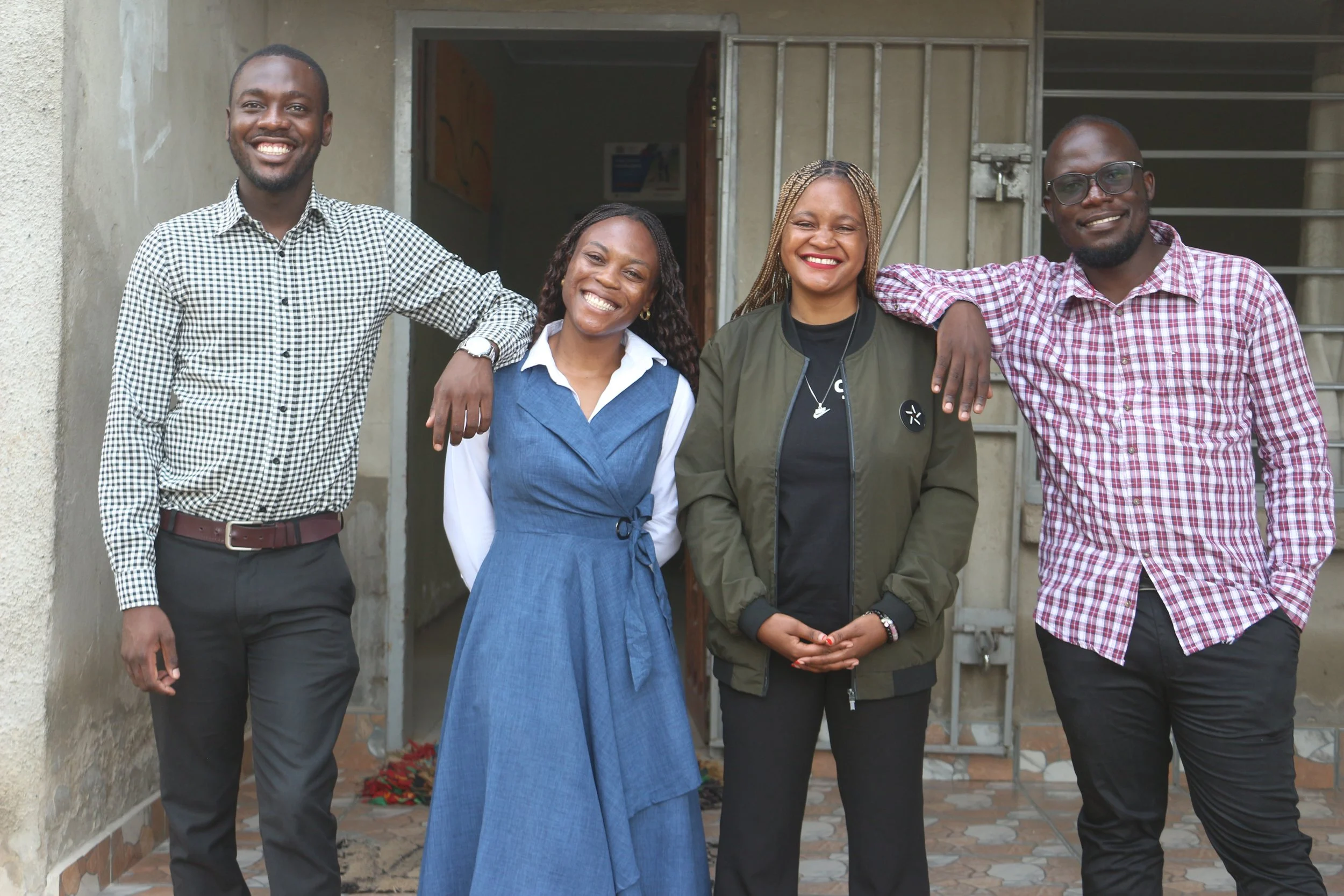 Two Zambian young women and two Zambian young men pose in front of their building and smile for the camera.