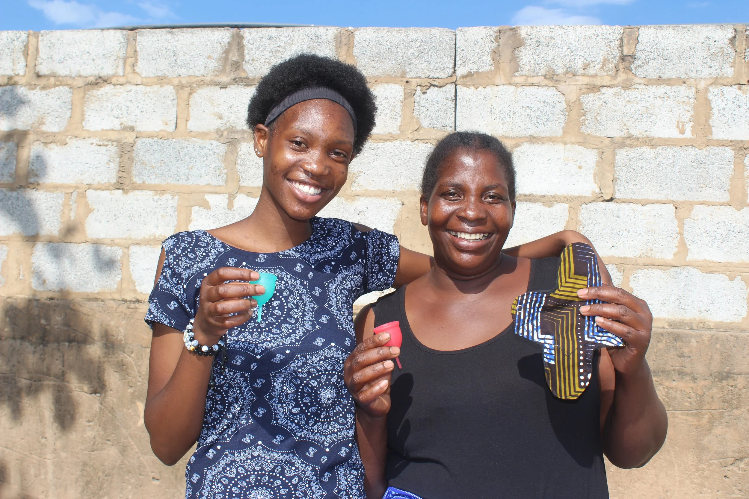 A young Zambian girl wearing a blue shirt and her mother, wearing a black shirt, are holding up menstrual cups and a menstrual pad and smiling while posing for the camera.