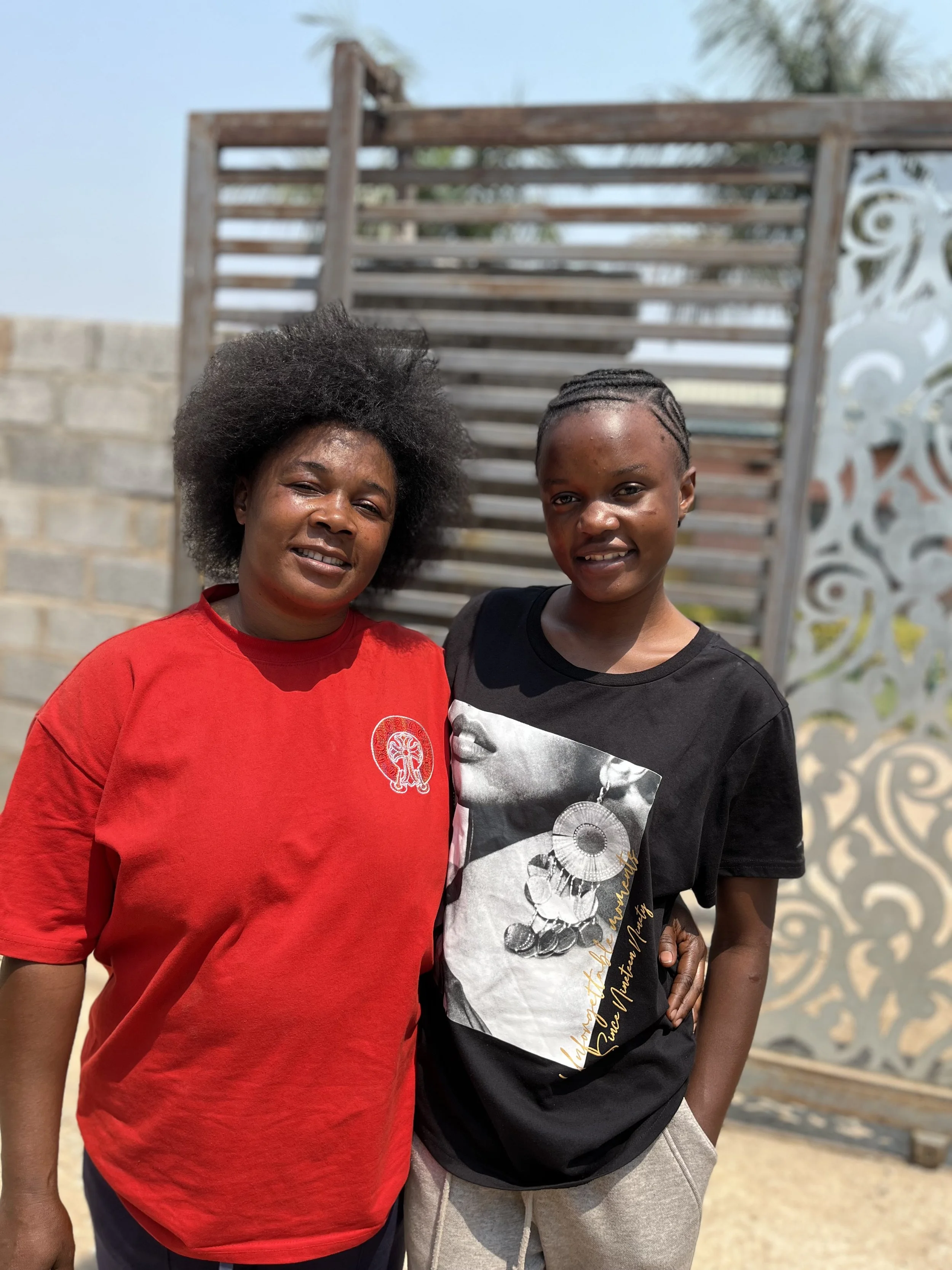 A Zambian mother wearing a red shirt puts her arm around her daughter who is wearing a black graphic shirt.
