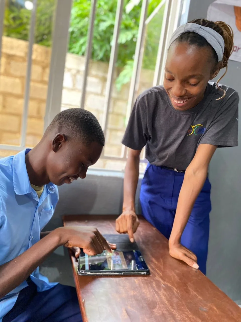 A young student wearing a blue shirt tries out a digital tablet while being directed by his teacher, a young Zambian woman wearing a grey tshirt and blue pants.