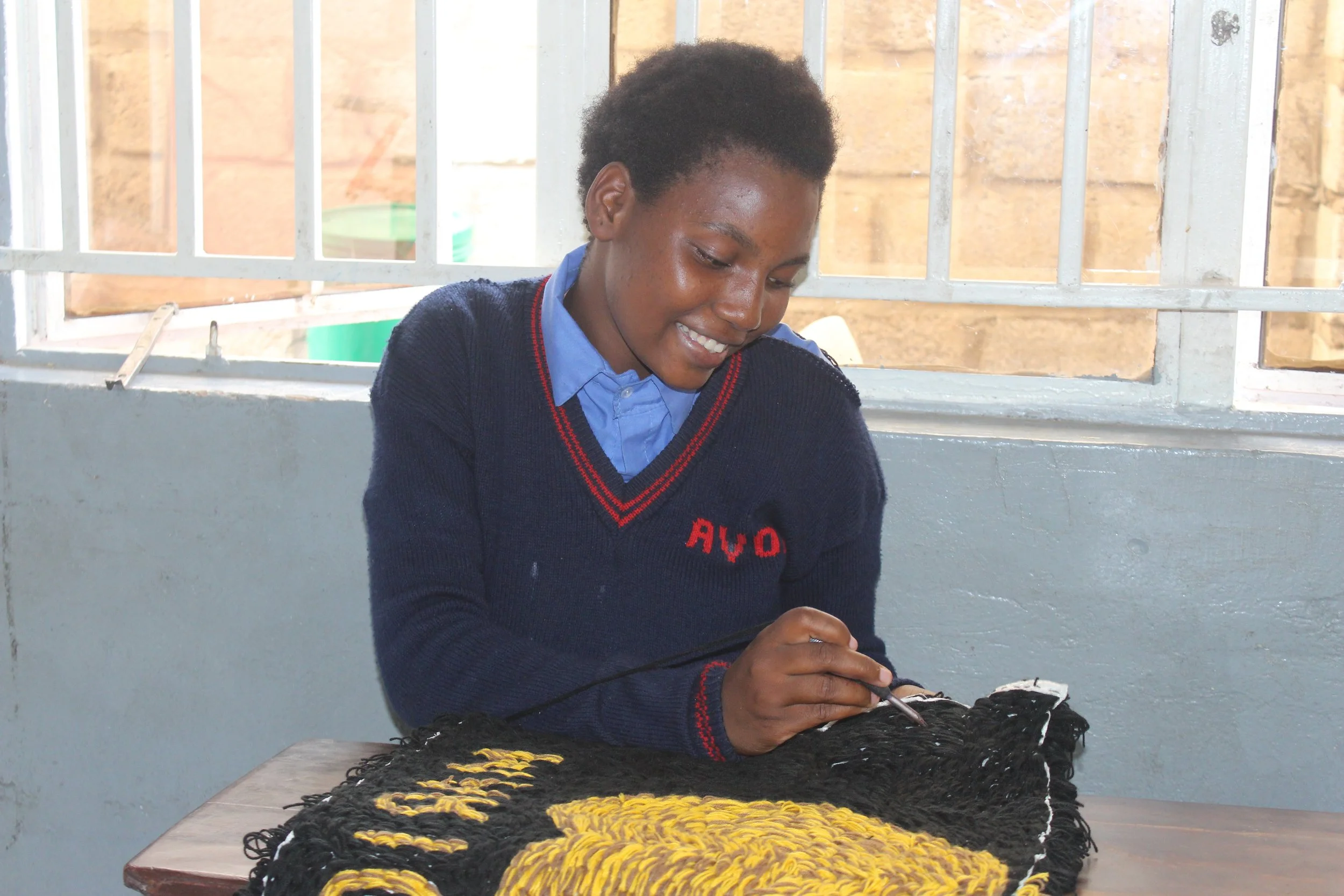 A young Zambian girl wearing a blue sweater knits a doormat.