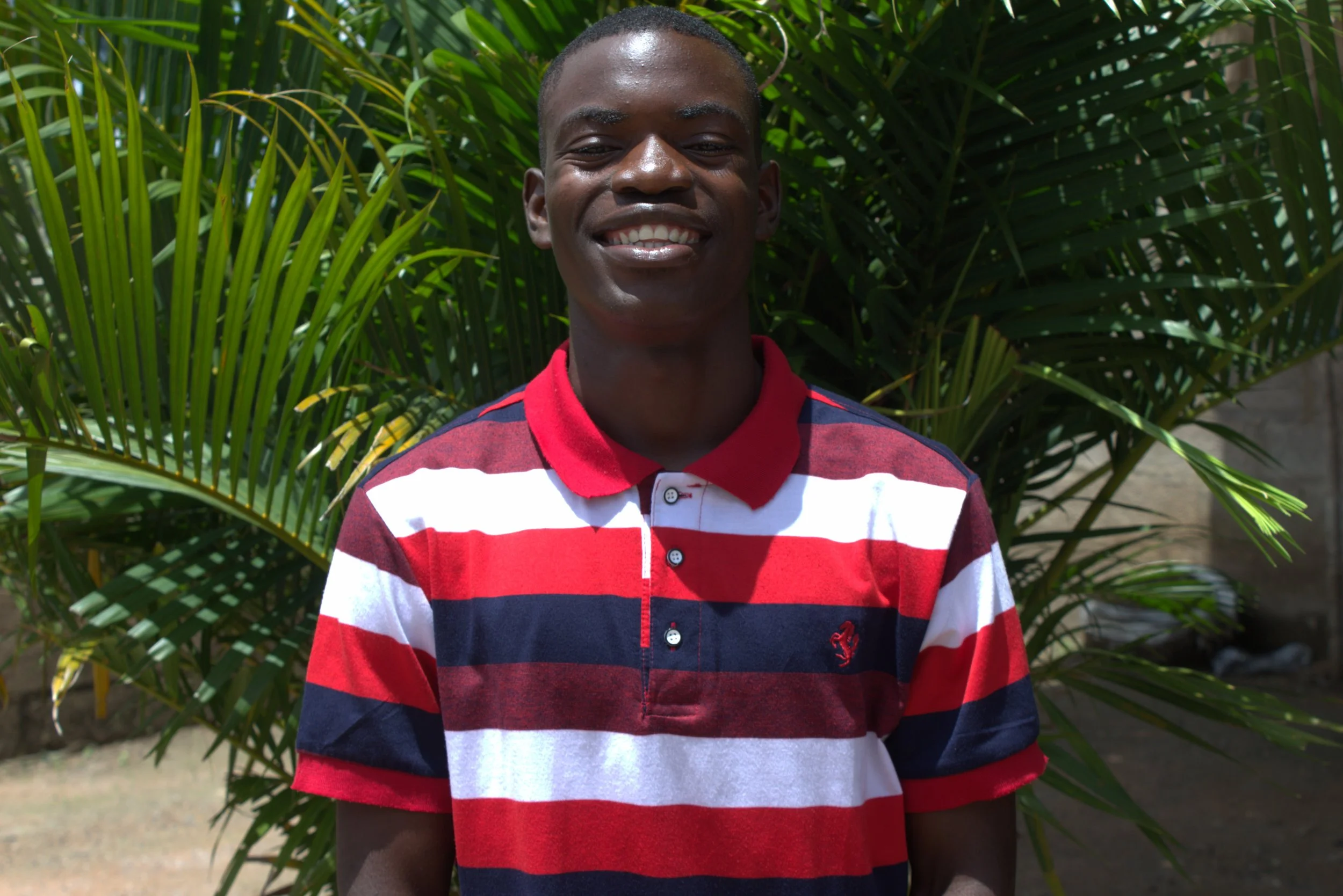 A young Zambian man wearing a red, white and blue striped shirt smiles for the camera.