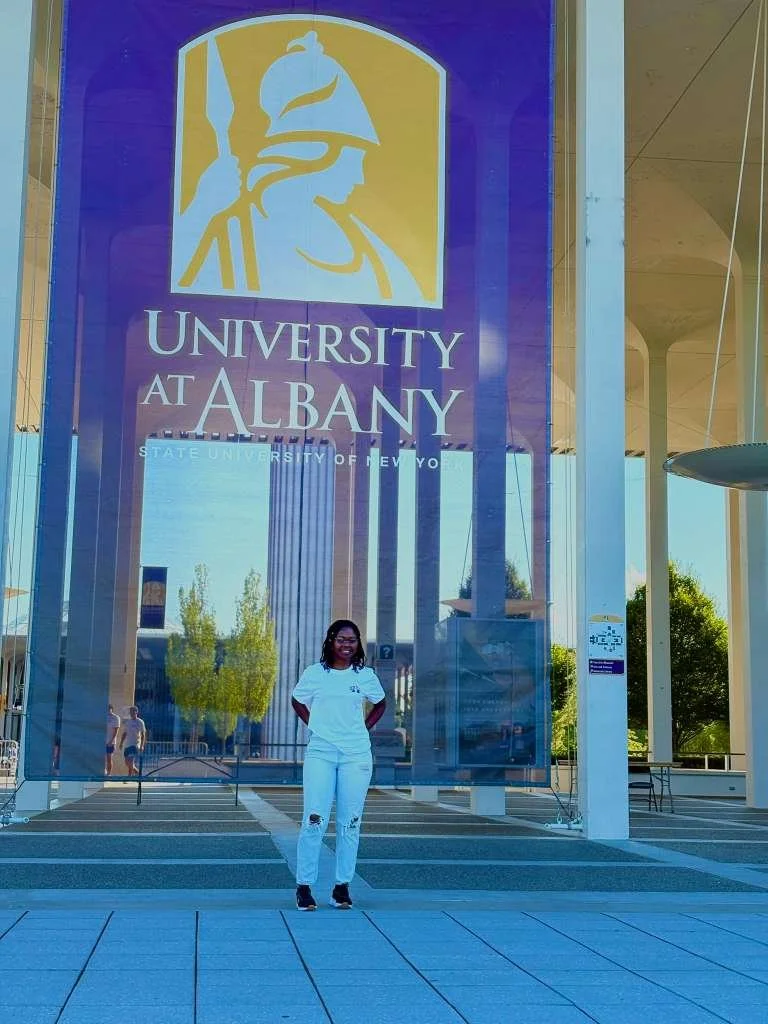 A Zambian college student stands in front of the University of Albany sign