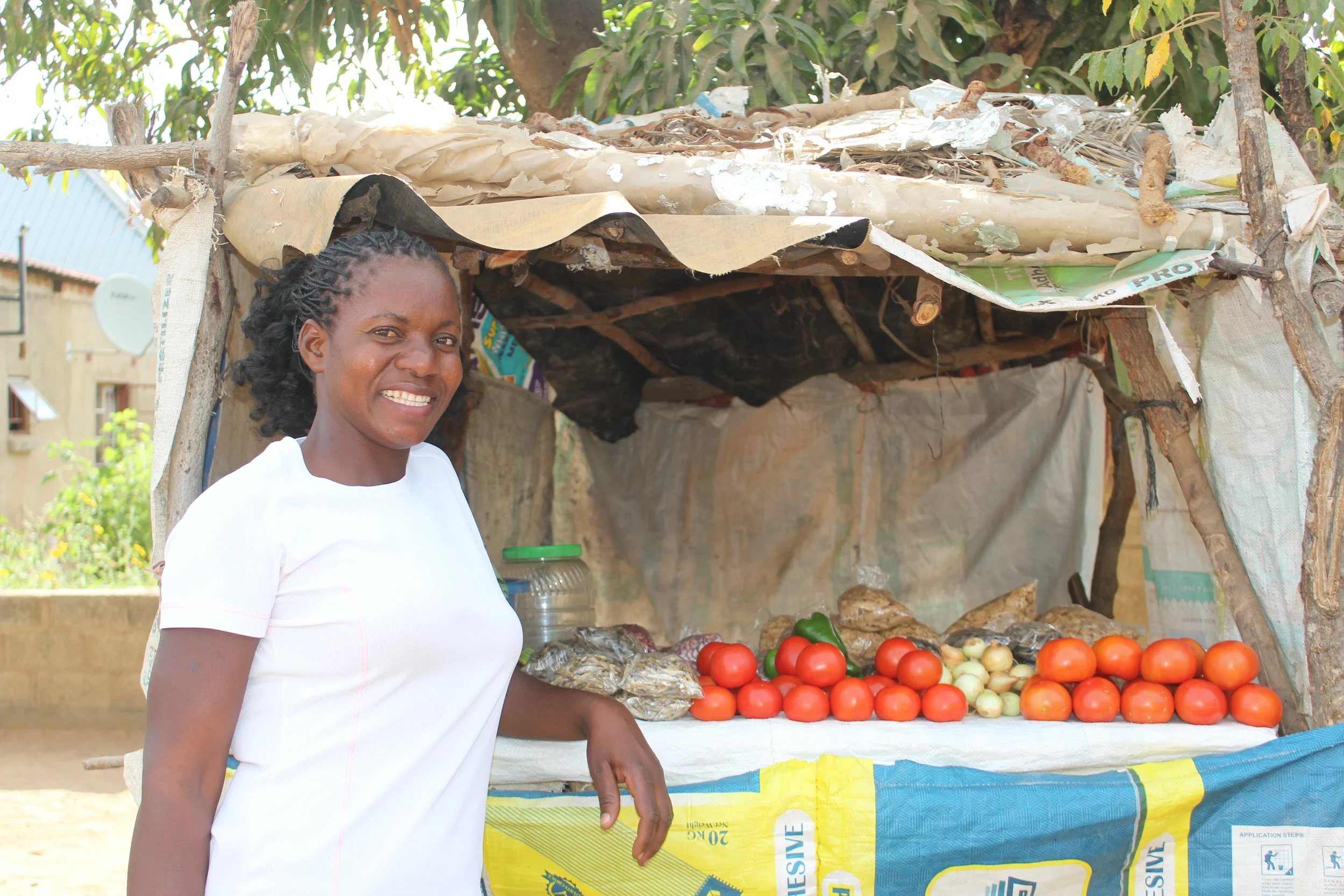 A Zambian woman wearing a white shirt smiles in front of her market stand showing red tomatoes.