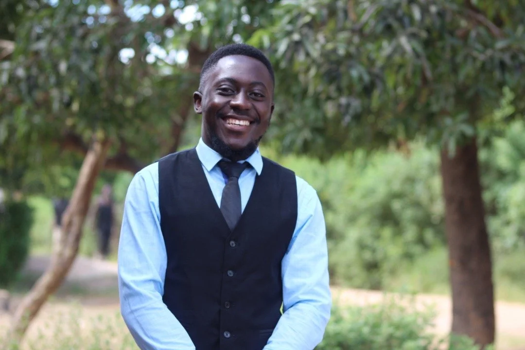 Photo of a Zambian male, Ceasar Samwaka, smiling for the camera wearing a blue shirt, vest and tie.