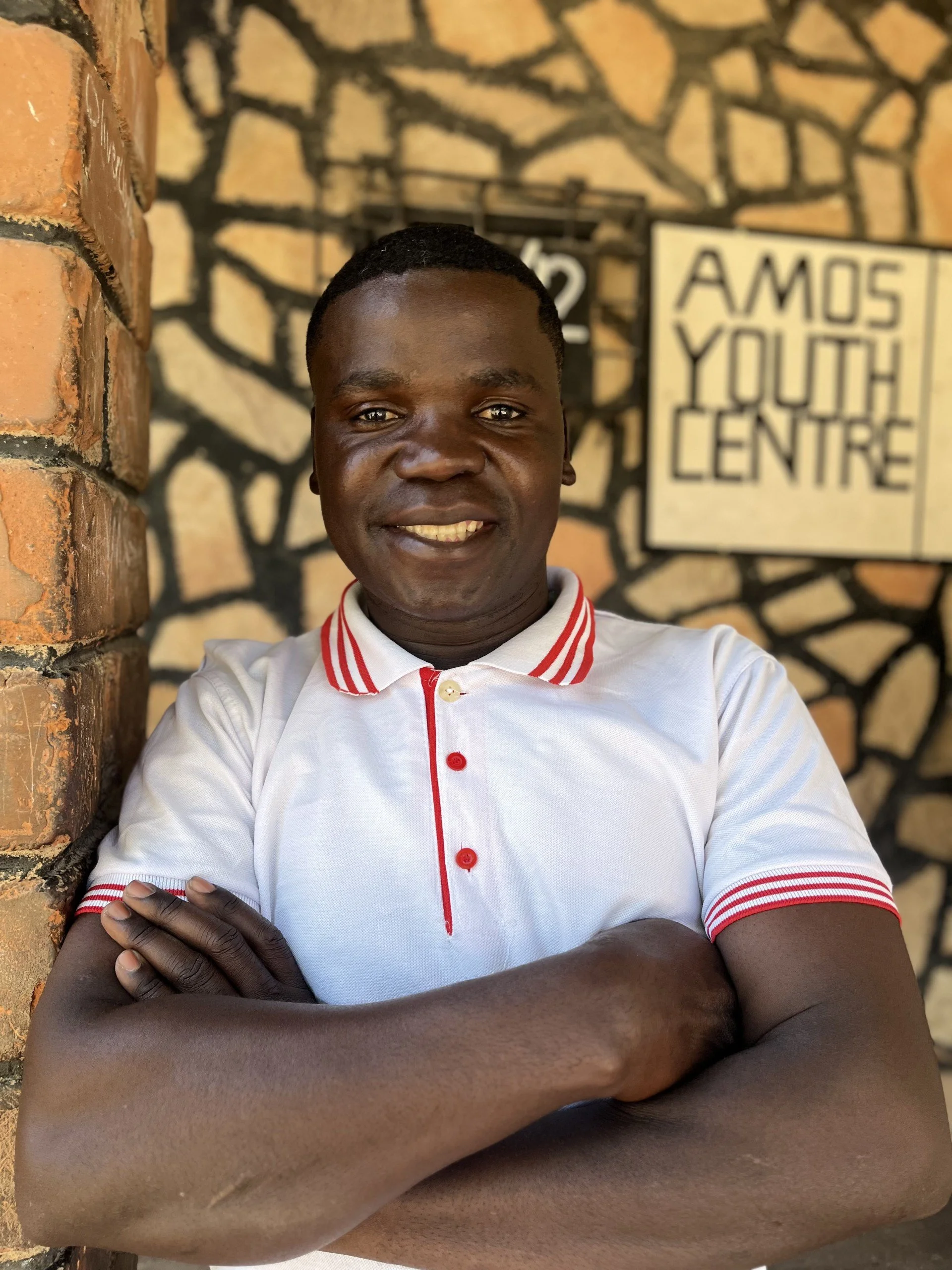A Zambian man wearing a white shirt with red trim is crossing his arms and smiling for the camera.