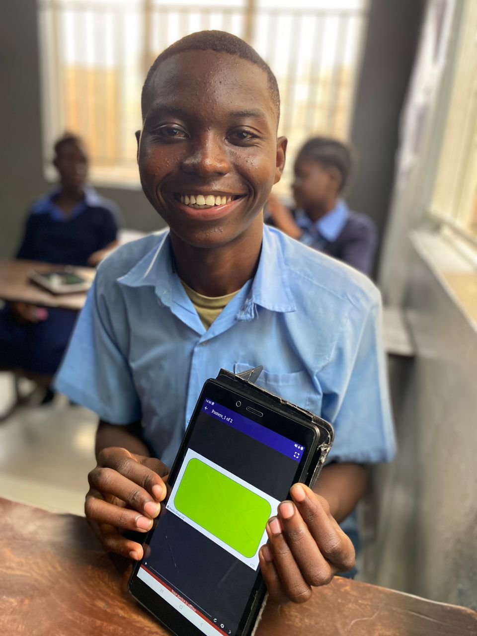 A young Zambian boy wearing a blue shirt and holding a tablet computer smiles for the camera.