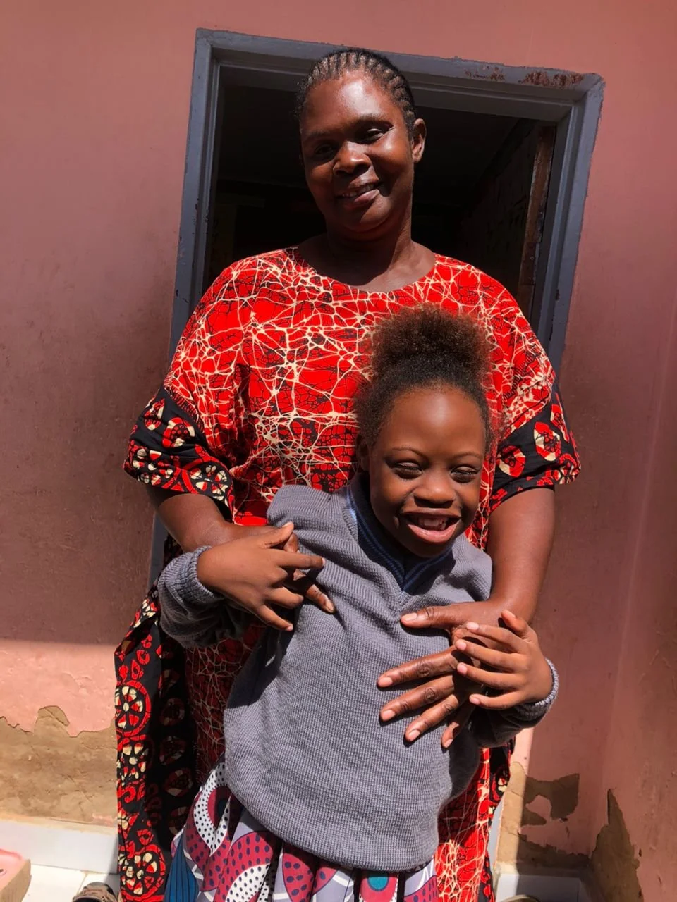 A Zambian mother wearing a red dress stands behind her daughter who has special needs. Hillary is wearing a blue long sleeve shirt, a colorful skirt and a big smile.