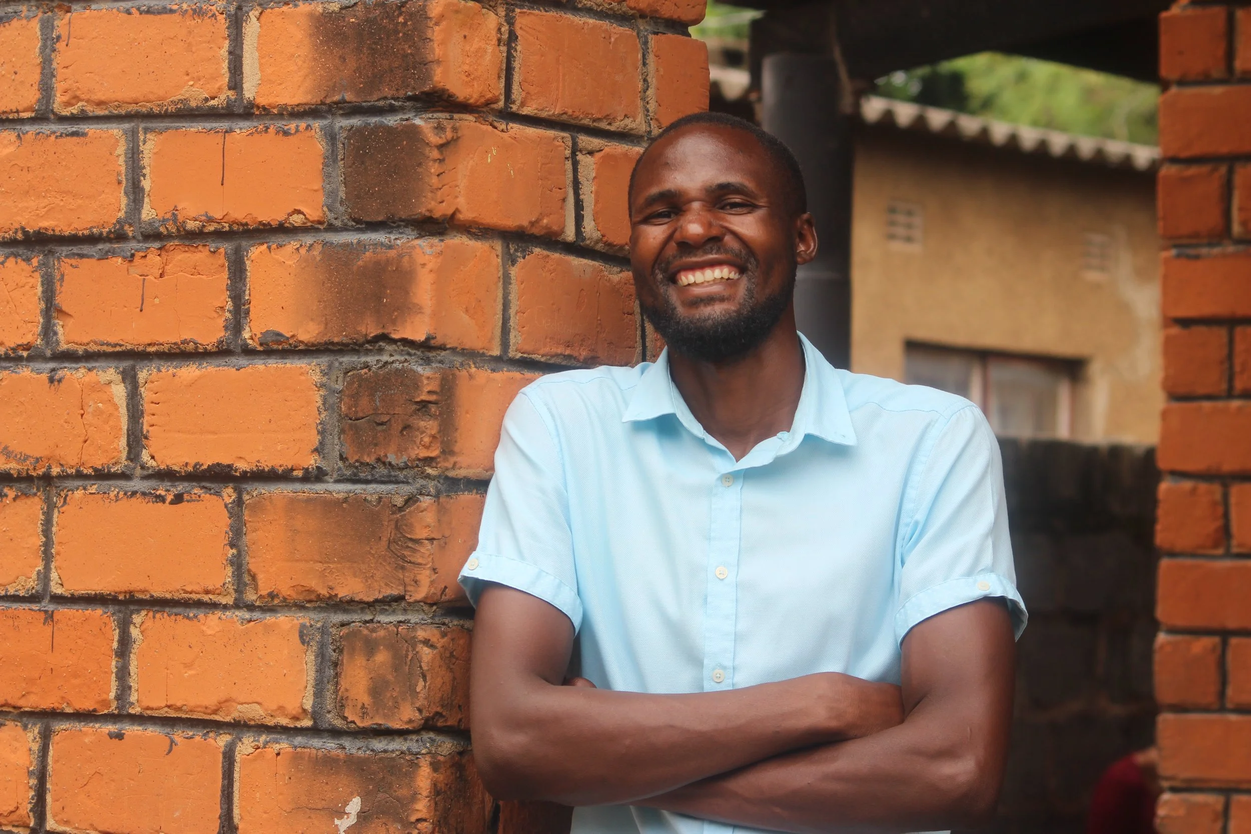 A young Zambian man is wearing a light colored polo shirt and smiling for the camera in front of a brick wall.