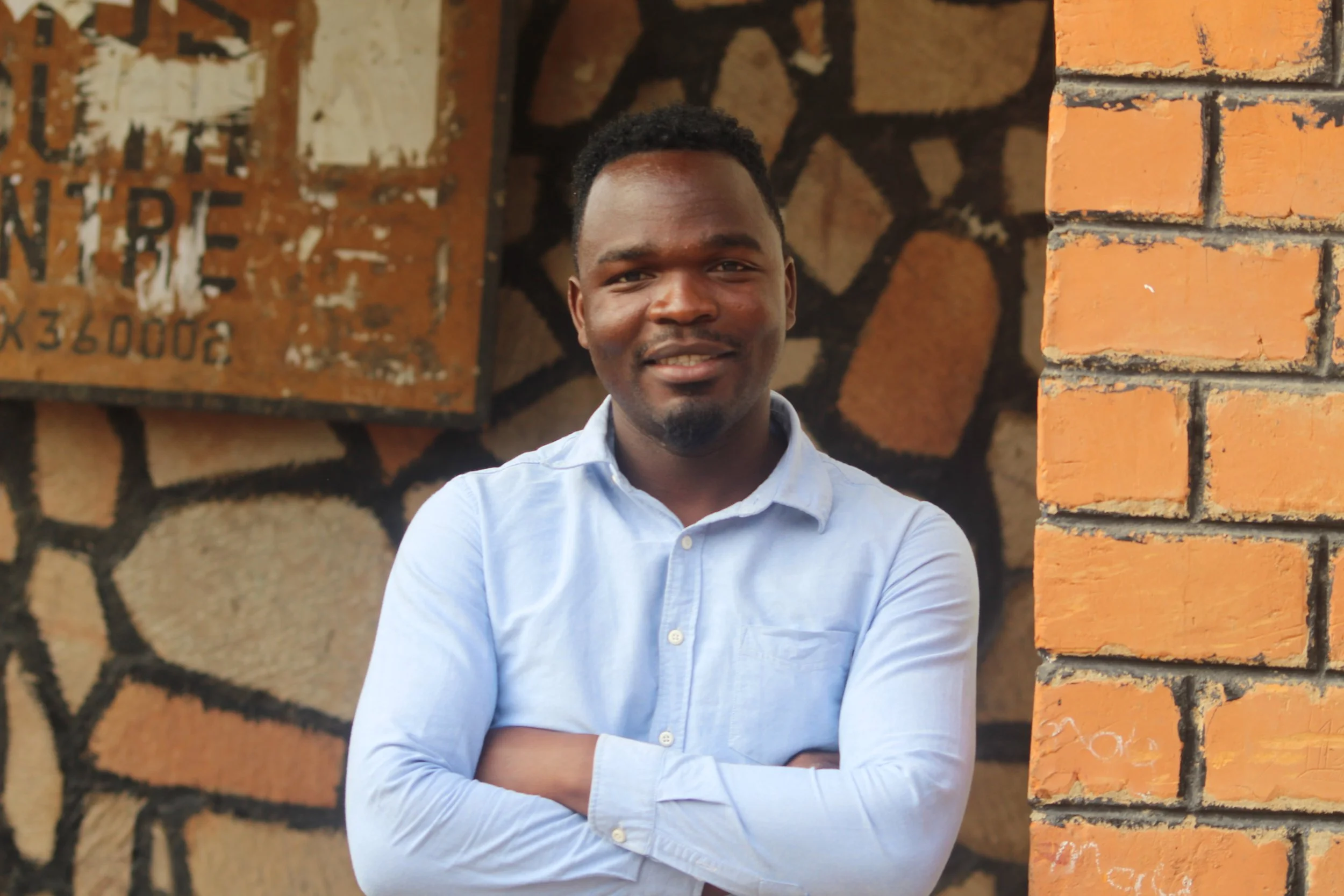 A Zambian man in his early 20s is wearing a light blue shirt and folding his arms while looking at the camera.