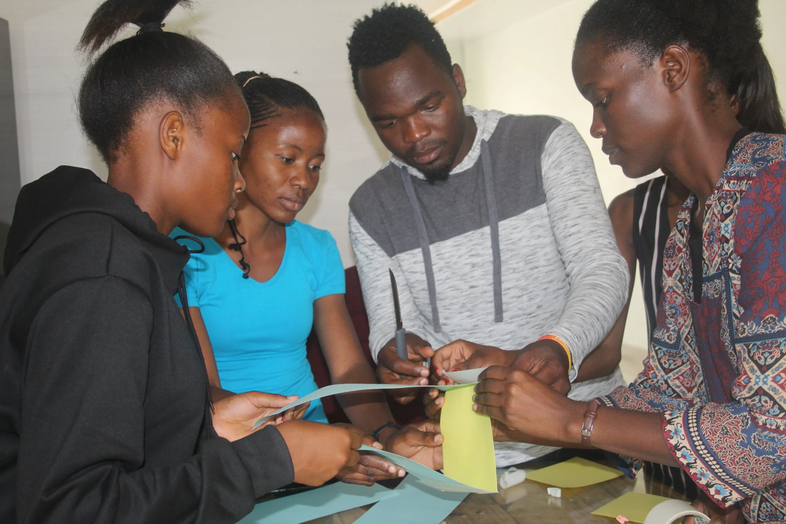 A Zambian male teacher wearing a grey sweatshirt is surrounded by three female students while working on a project in the classroom.
