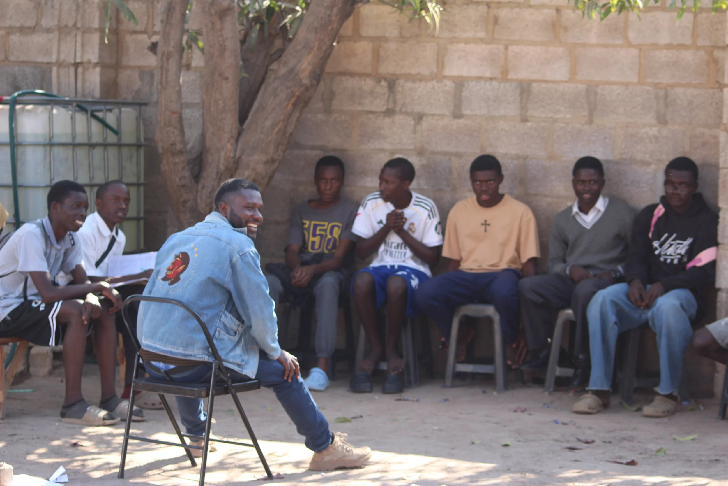 A Zambian teacher wearing a jean jacket and blue pants sits on a brown chair in front of a group of boys ready to lead a club.