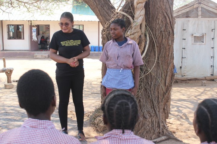 A Zambian young woman wearing a black t-shirt and black jeans stands in front of her students outside under a tree.