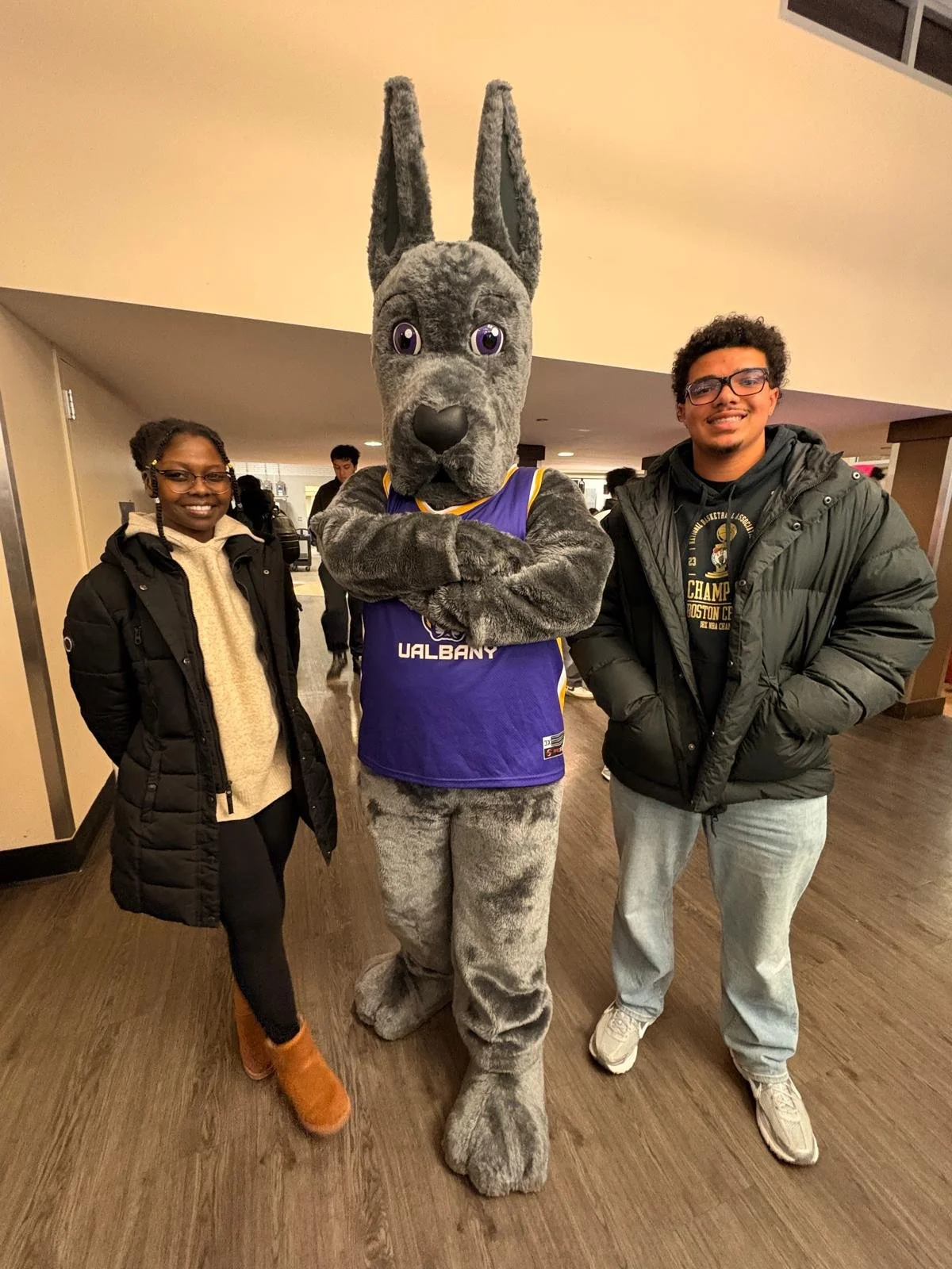 Two college students stand next to the University of Albany mascot, a large grey dog.
