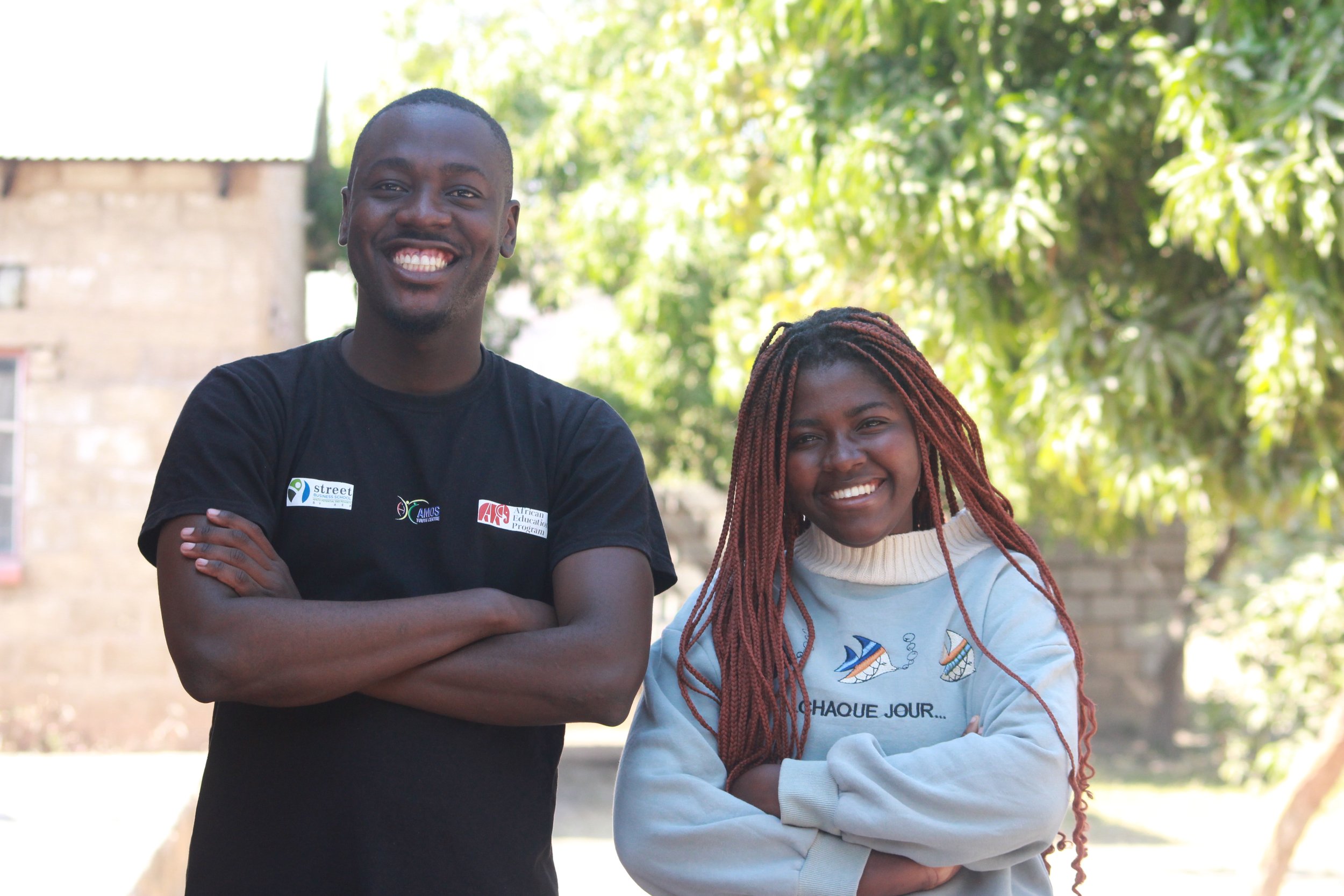A Zambian girl wearing a blue sweatshirt and her hair in braids stands next to a Zambian man wearing a black shirt, both smiling for the camera.
