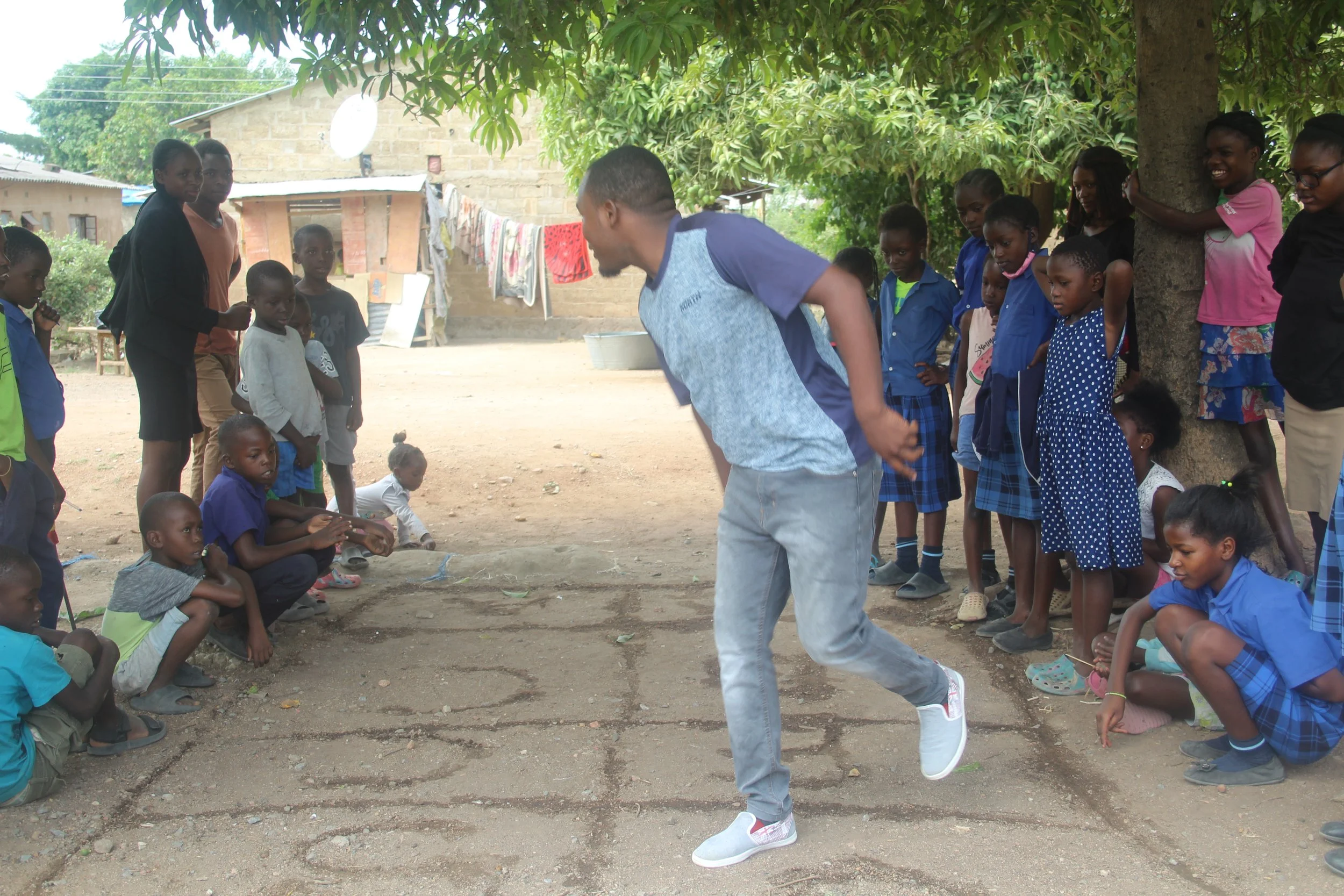 A male Zambian teacher wearing a blue shirt and blue jeans plays native games to teach Young Learners at AYC.