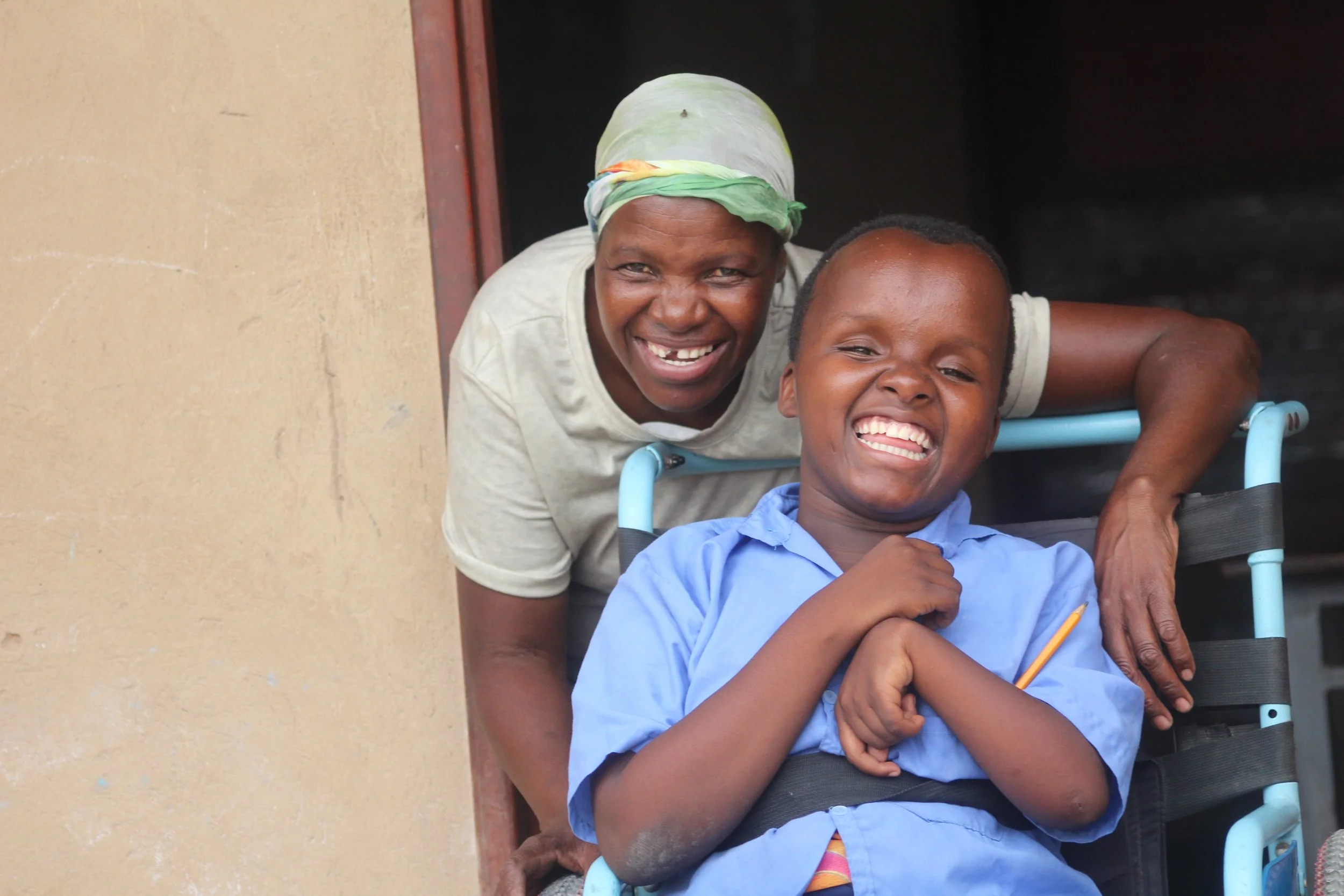A Zambian mother wearing a head scarf leans over her daughter in her wheelchair as they both smile for the camera.