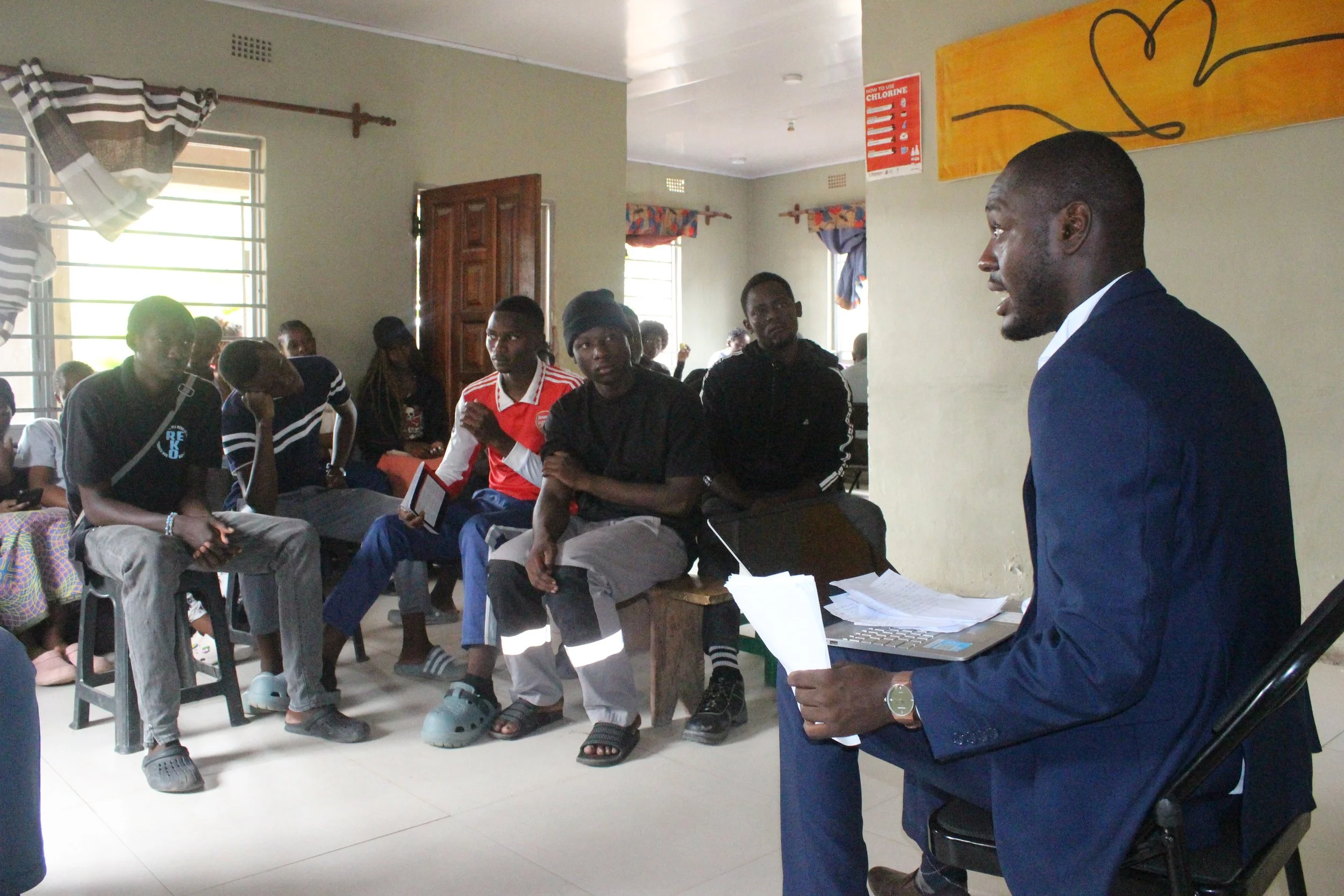 A Zambian young man wearing a blue suit is a teacher addressing his male Zambian students in a classroom.