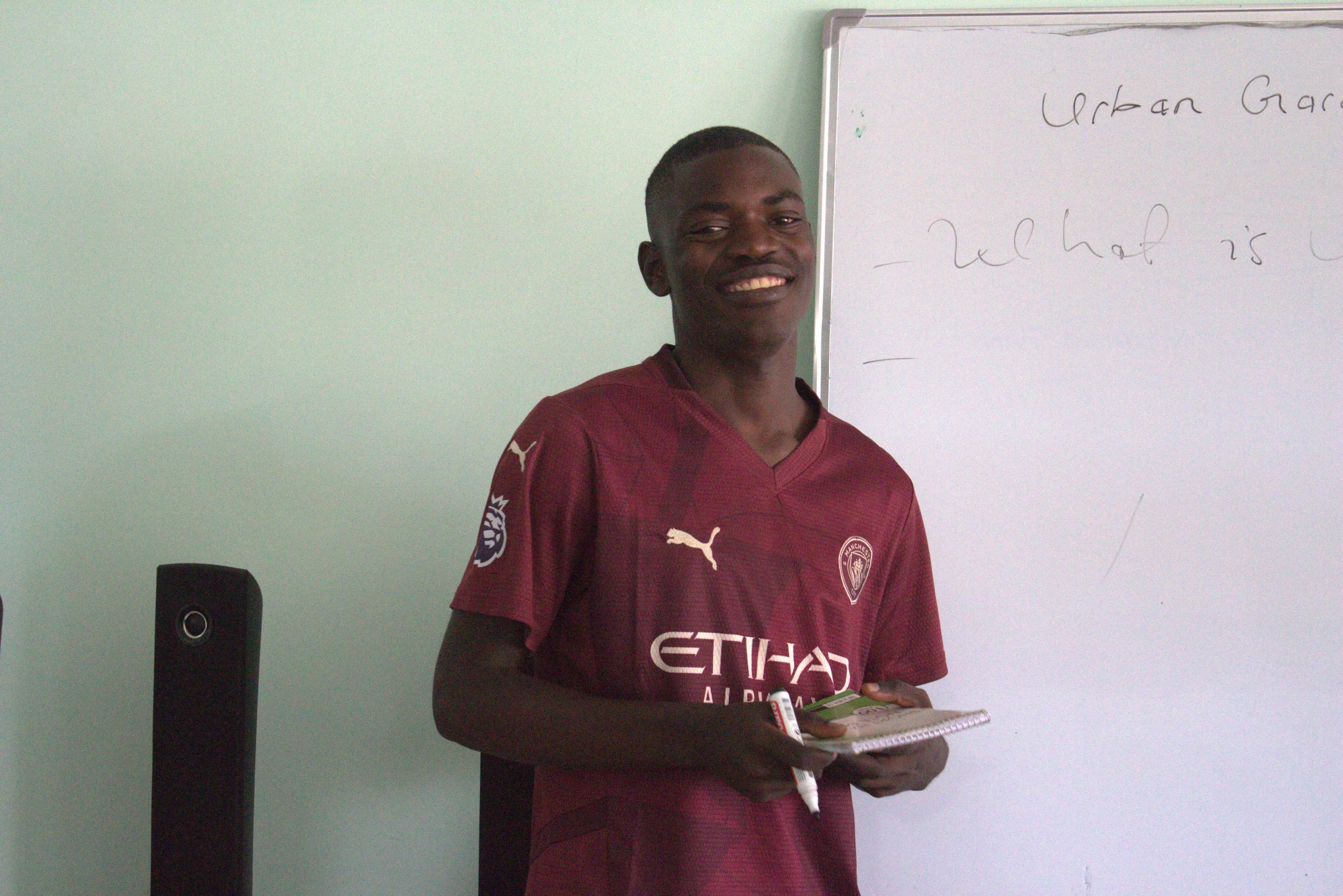 A young Zambian man wearing a red sports shirt stands in front of the classroom teaching his peers.