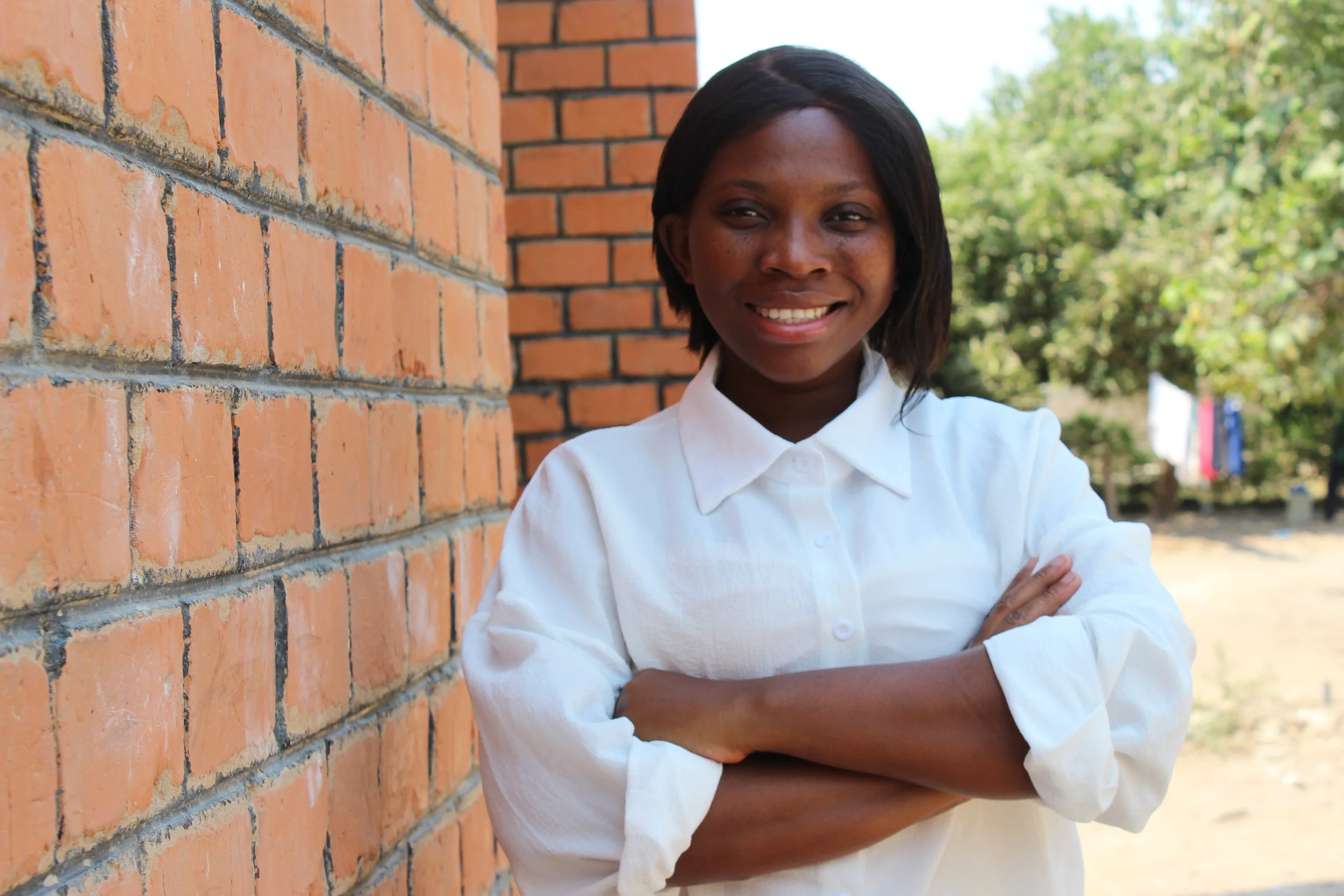 A Zambian woman wearing a white shirt is standing in front of a brick wall with her arms crossed.