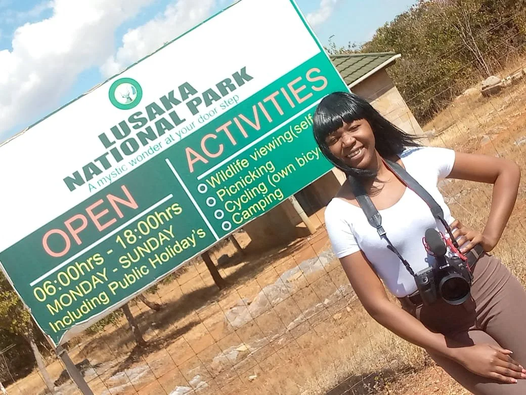 Zambian women wearing a white top stands in front of the Lusaka National Park entrance sign while holding a camera.