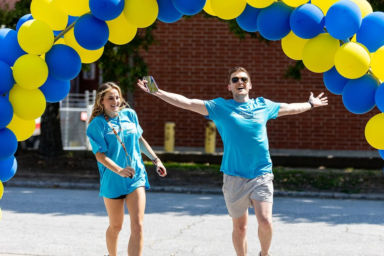 Two Inspiredu event participants in blue shirts celebrate beneath a blue and yellow balloon arch.