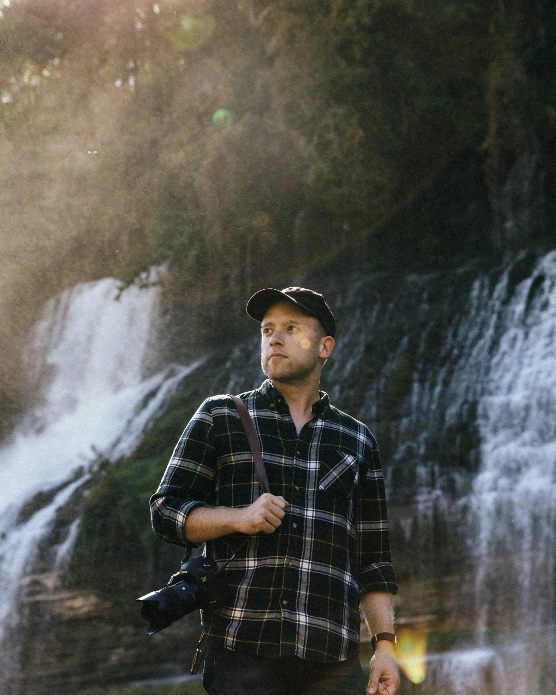 Jared Whitney, photographer and real estate agent, casually stands in front of a waterfall with a plaid shirt.