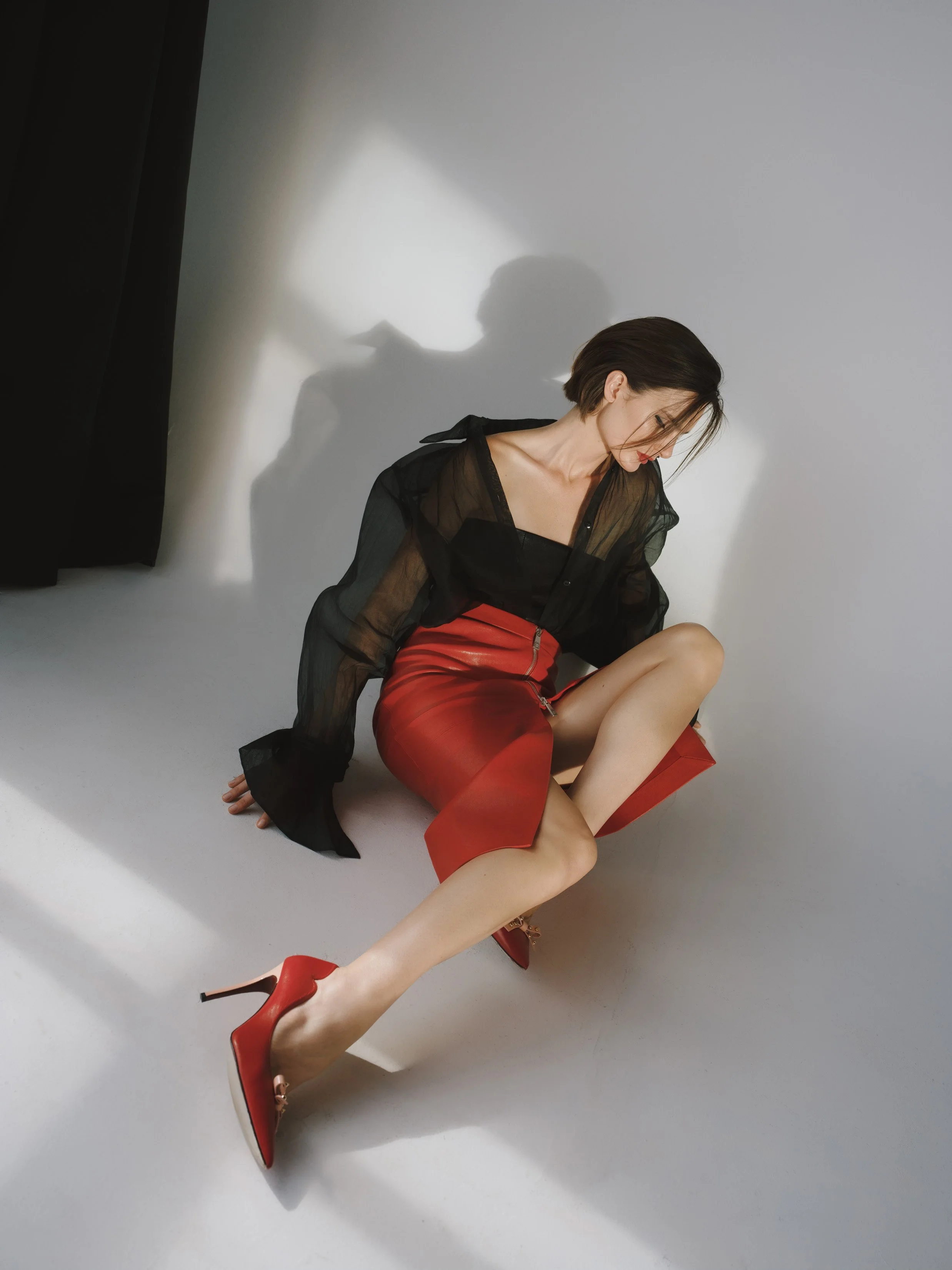 A woman with short dark hair is sitting on the floor, wearing a sheer black top, a red skirt with a zipper, and red high heels. She is looking downward, and her shadow is cast on the white wall behind her.