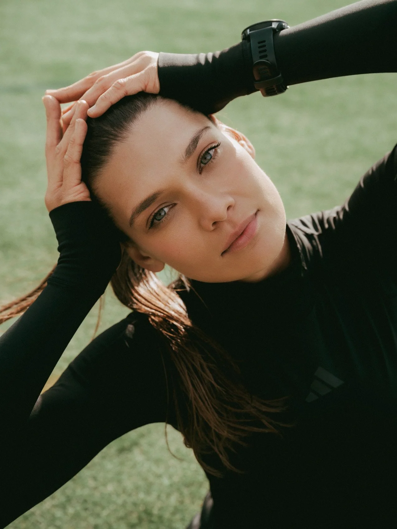 A young woman with long brown hair, wearing a black sporty outfit, lying on a grassy field, looking into the camera, holding her head with her hands.