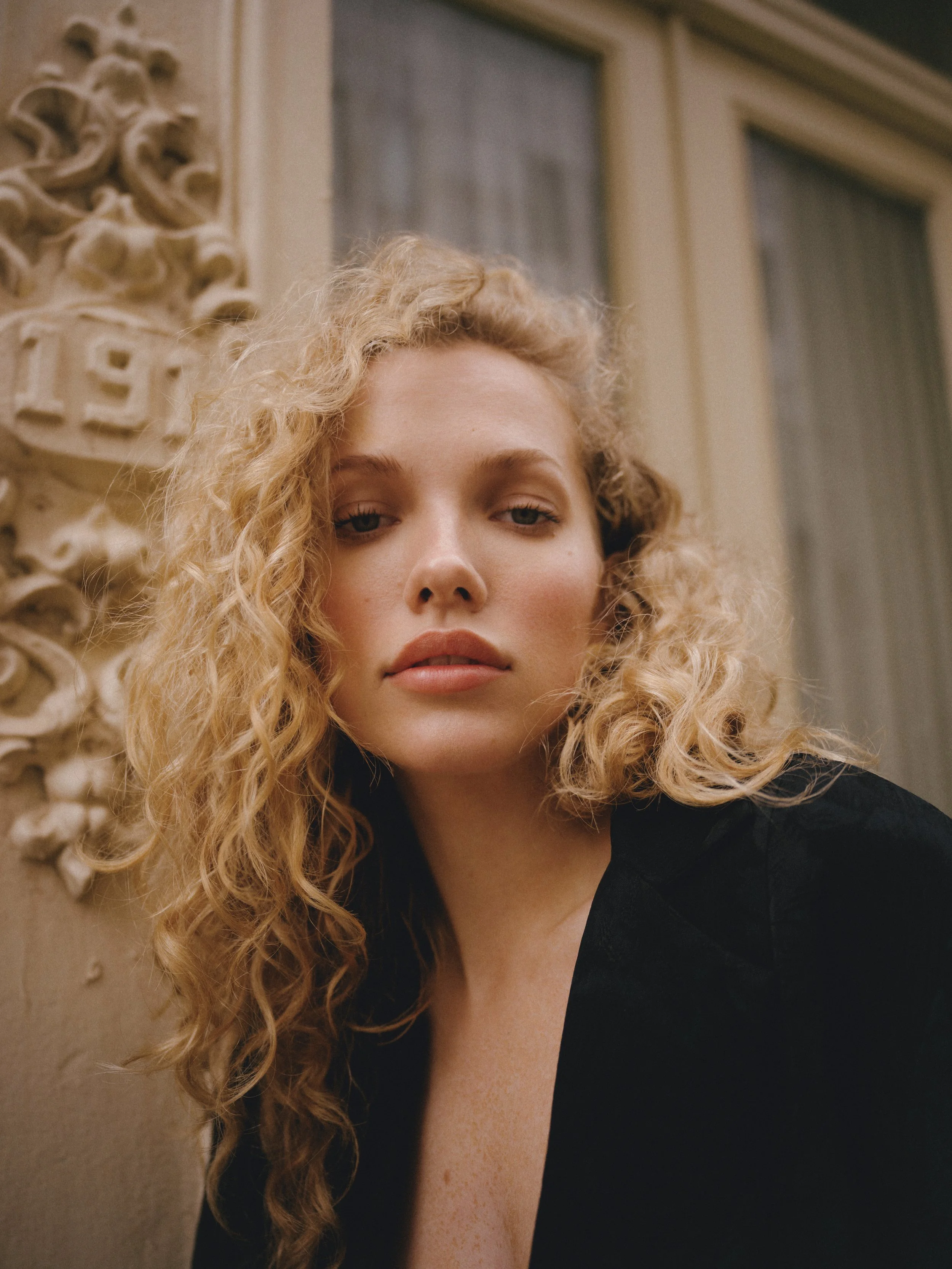 A close-up of a woman's face with curly blonde hair, standing outdoors near architectural details with ornate carvings, and two windows in the background.