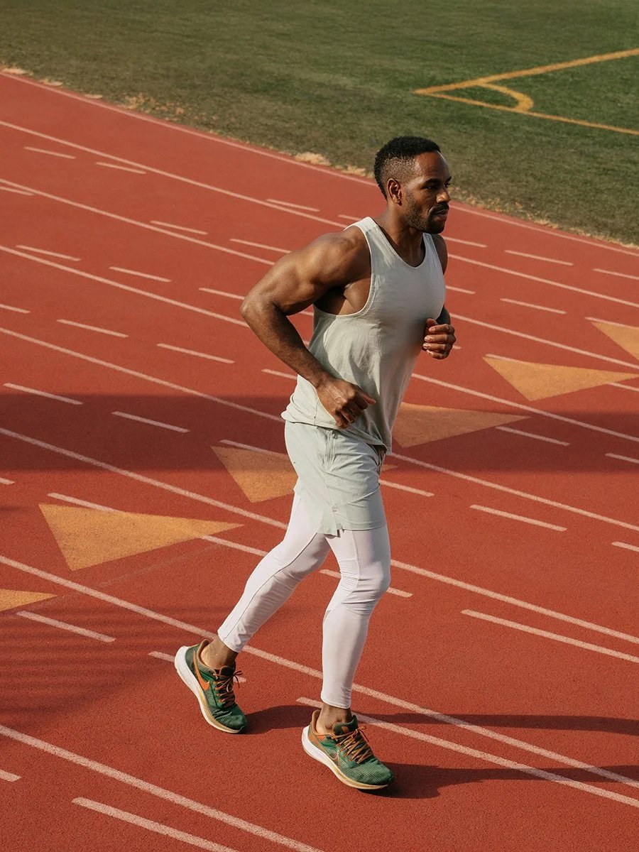 A man running on a red outdoor track during daytime, wearing a sleeveless gray athletic shirt, white leggings, and green running shoes.