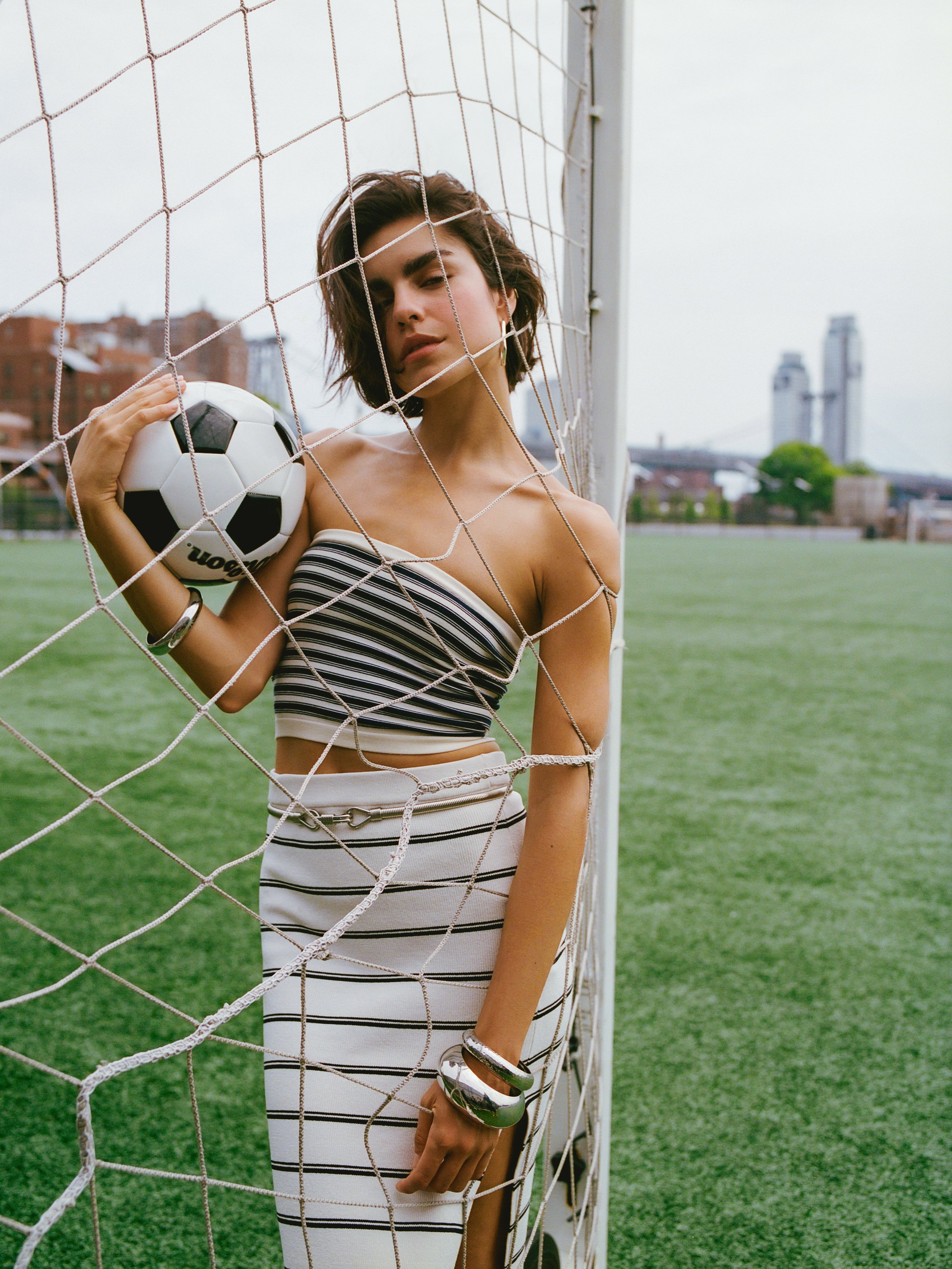 A woman standing in front of a soccer goal net on a field, holding a soccer ball, with a cityscape in the background.