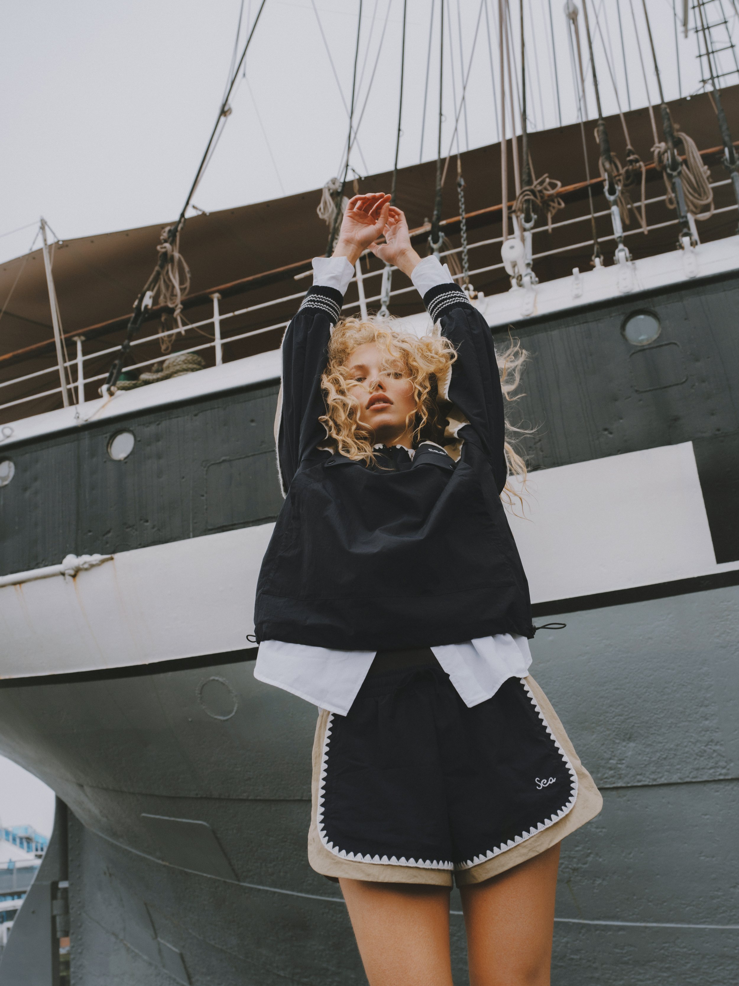 Young woman with curly blonde hair standing in front of a ship, wearing a black jacket, beige shorts, and a white shirt.