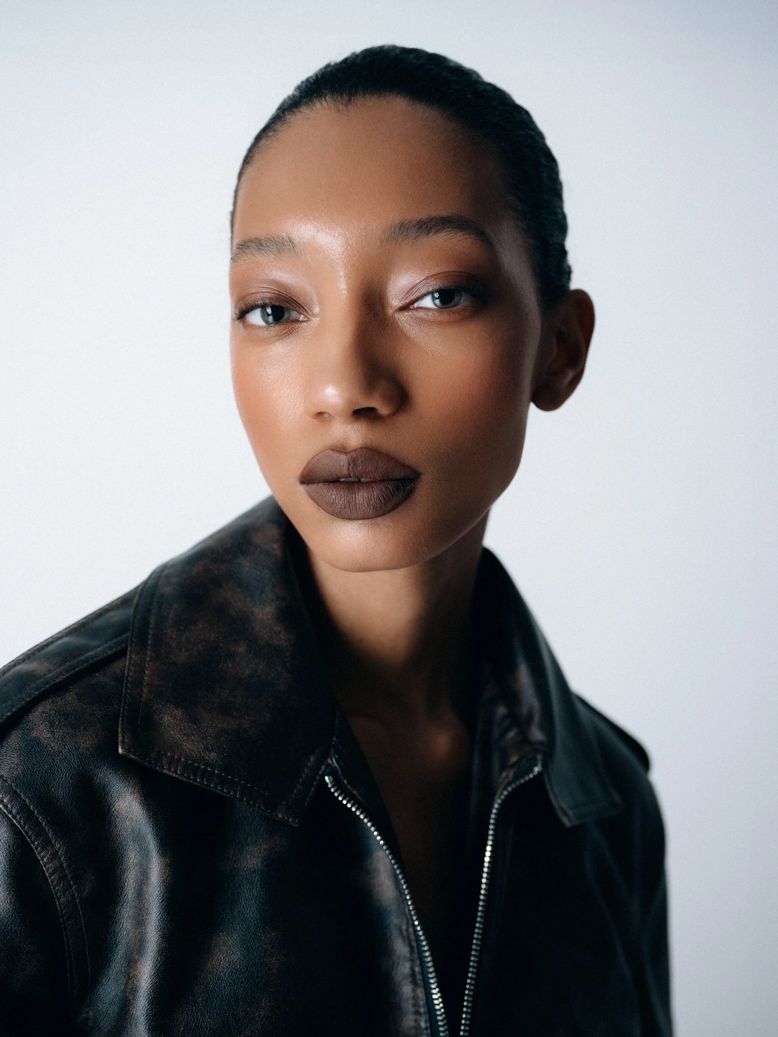 Close-up of a woman with dark skin, wearing a black leather jacket, looking directly at the camera with a neutral expression.