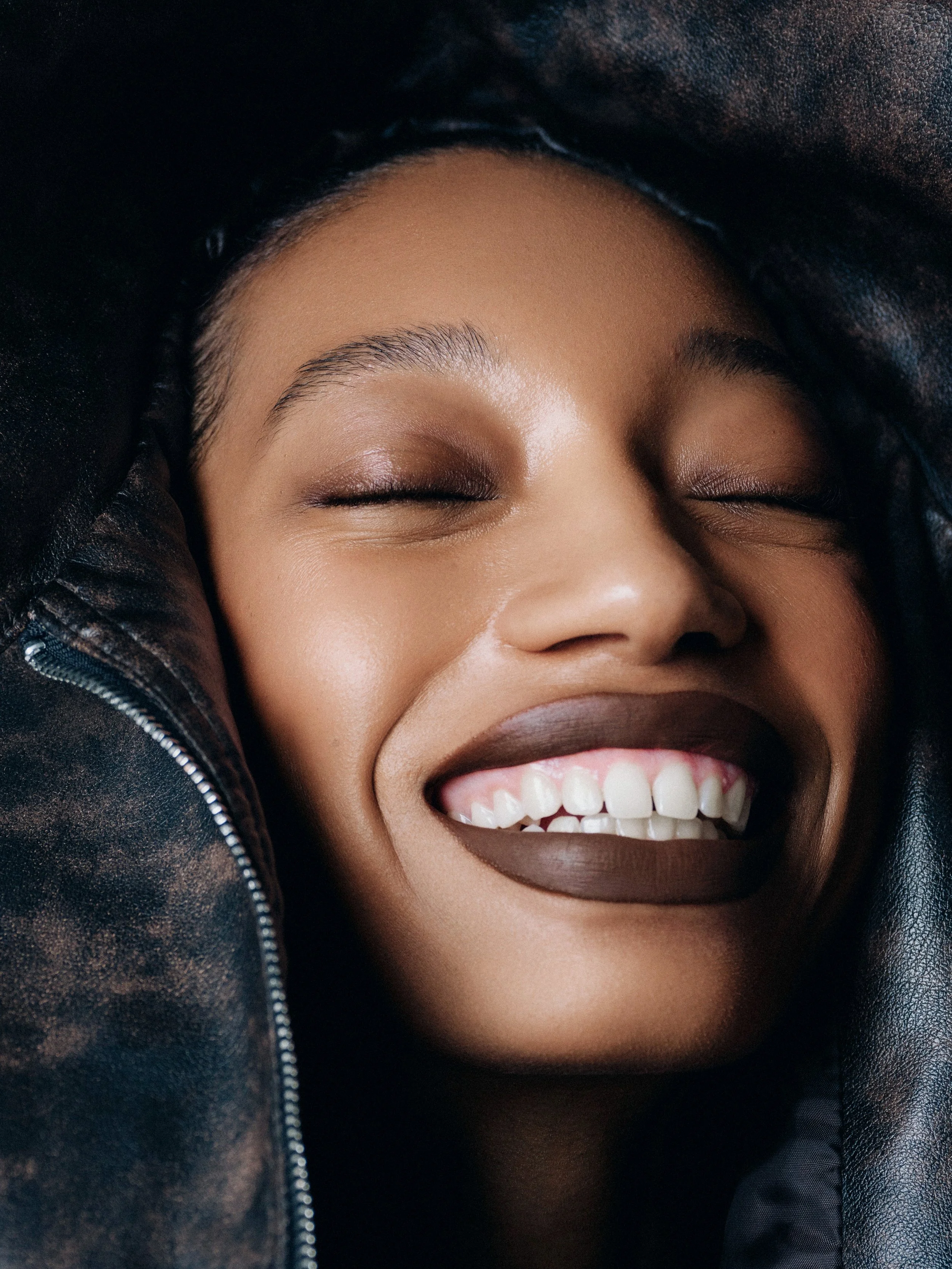 Close-up of a woman smiling with eyes closed, wearing dark lipstick and a black leather jacket with a zipper.