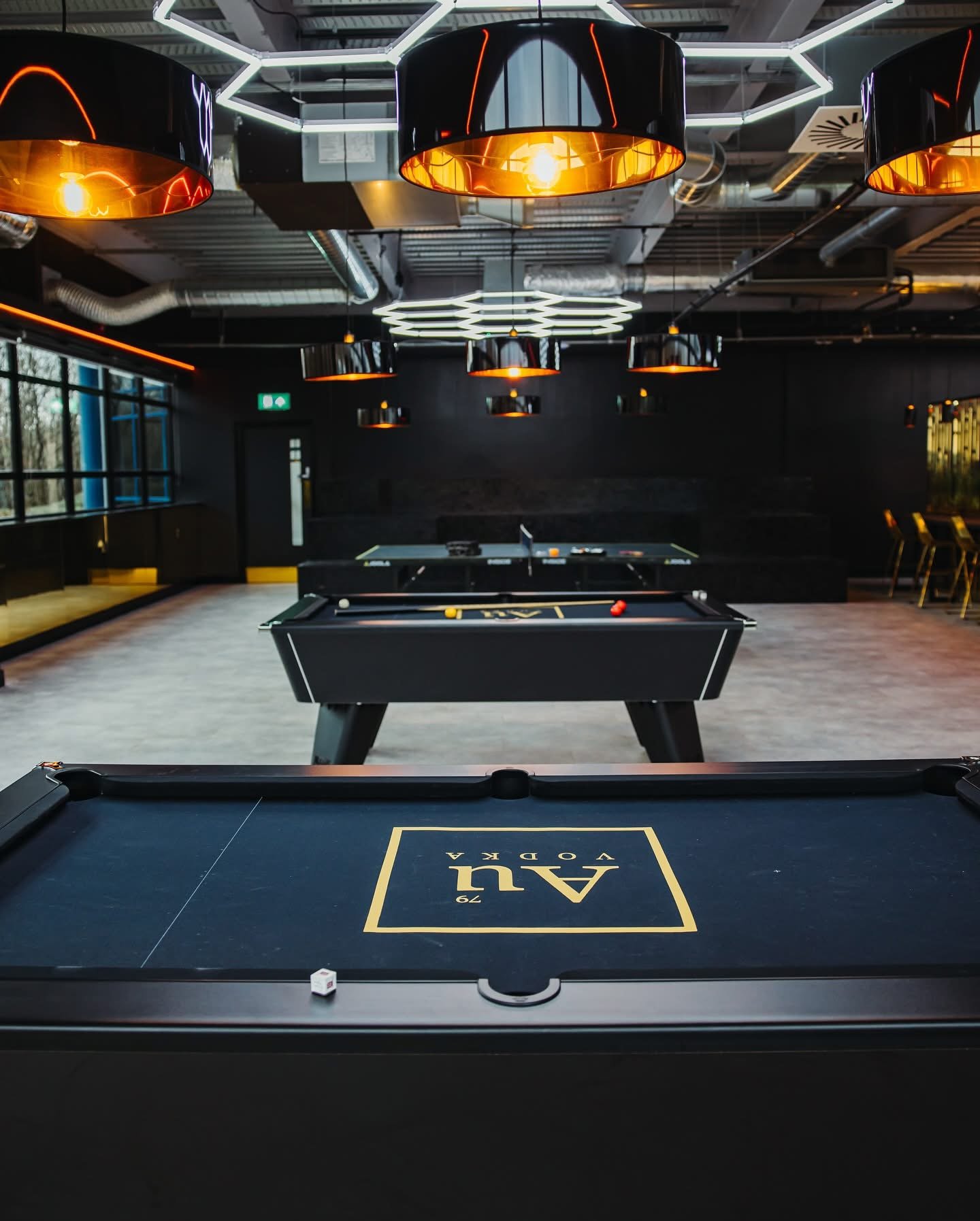 Interior of a modern game room with black ceiling and walls, featuring multiple hanging black and gold pendant lights, and black benches along the walls. In the center, there are two billiard tables, and in the foreground, a black table with the brand name 'AQUA' printed on it.