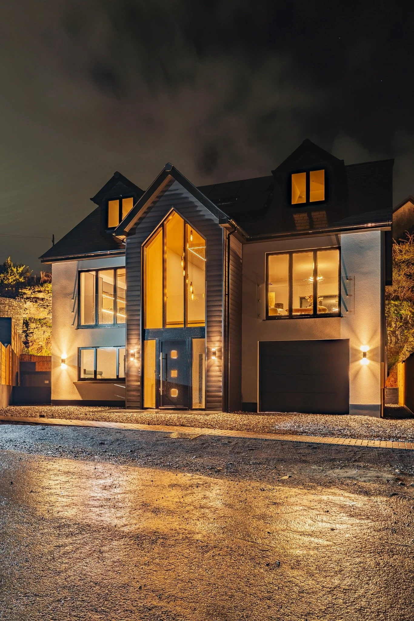 A modern, illuminated house at night with large glass windows and a black roof, situated on a wet driveway.
