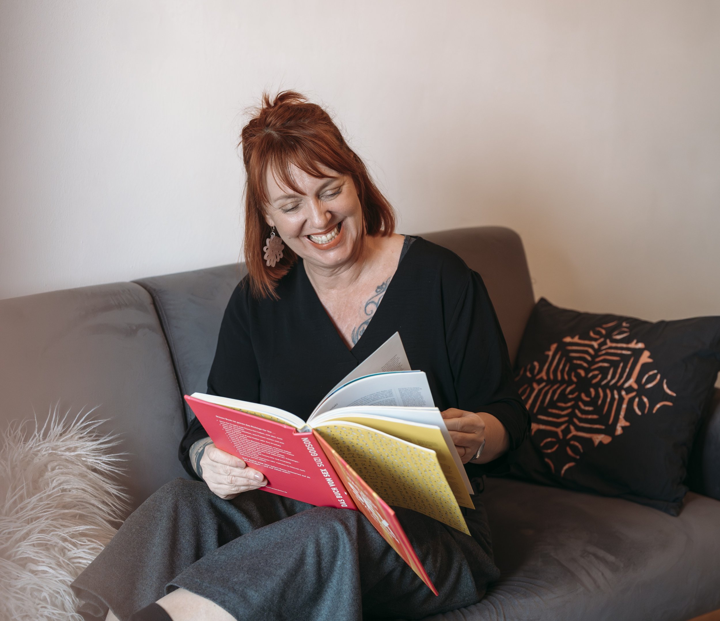 A woman with red hair, earrings, and tattoos is sitting on a gray couch, smiling and reading a magazine. She is dressed in a black top, and the background has a plain light-colored wall.