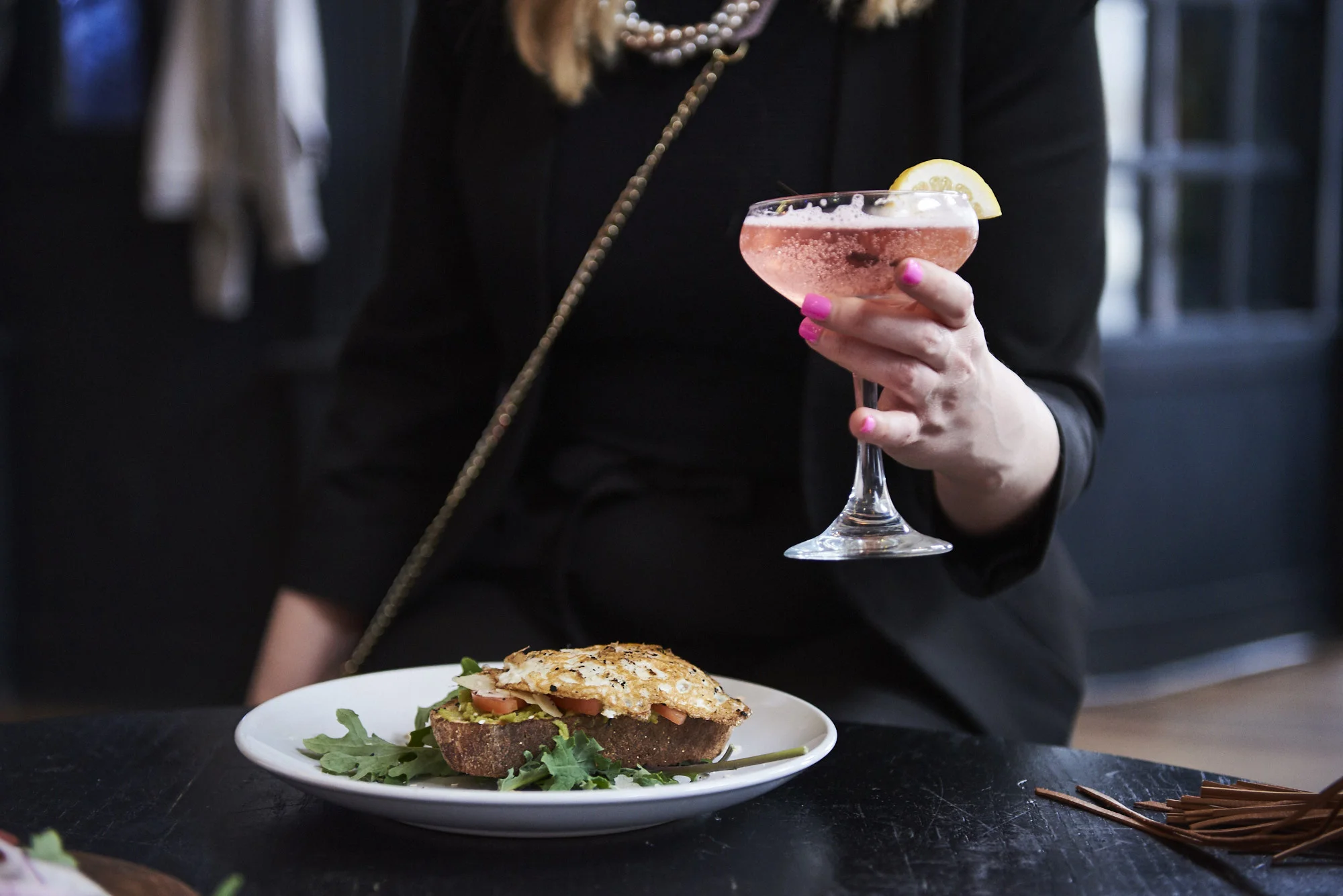 A woman holding a pink cocktail and a sandwich.