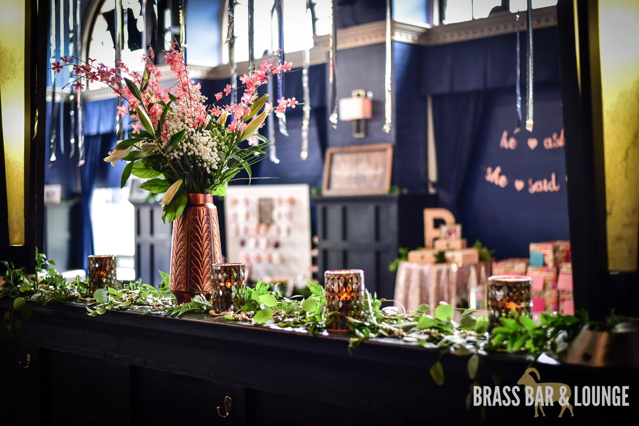 A ledge decorated with candles, green leaves, and a rose gold metallic vase with flowers.