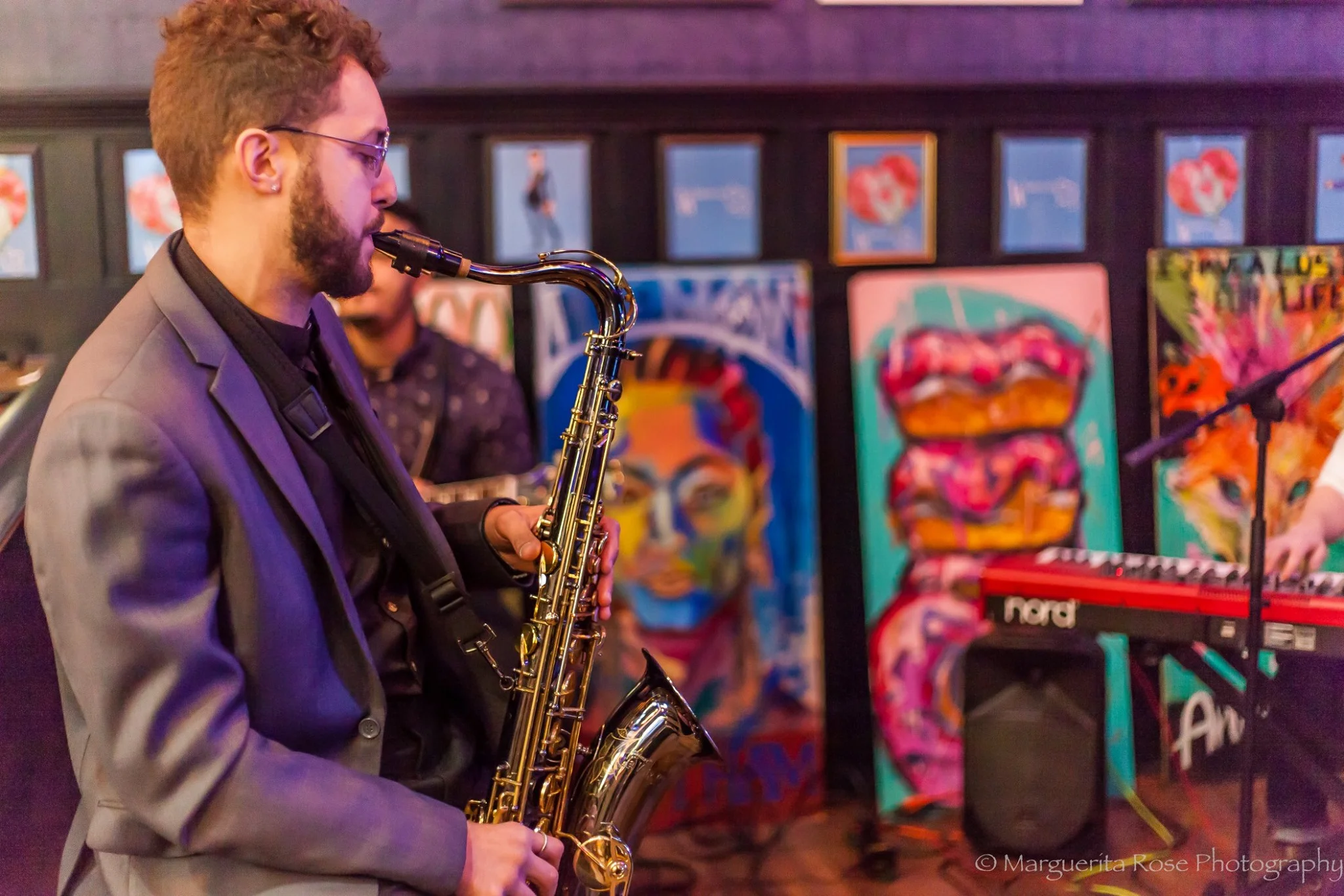 A man wearing glasses playing a saxophone with paintings and a keyboard in the background.