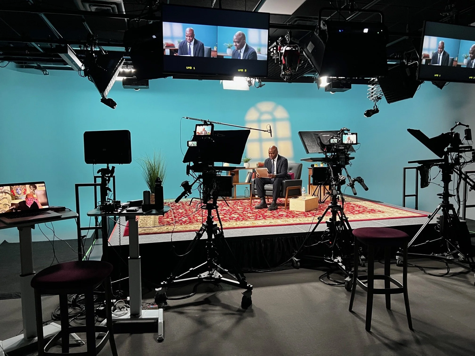 Behind-the-scenes view of a professional video production studio with cameras, lighting equipment, monitors, and a seated speaker on a turquoise set.