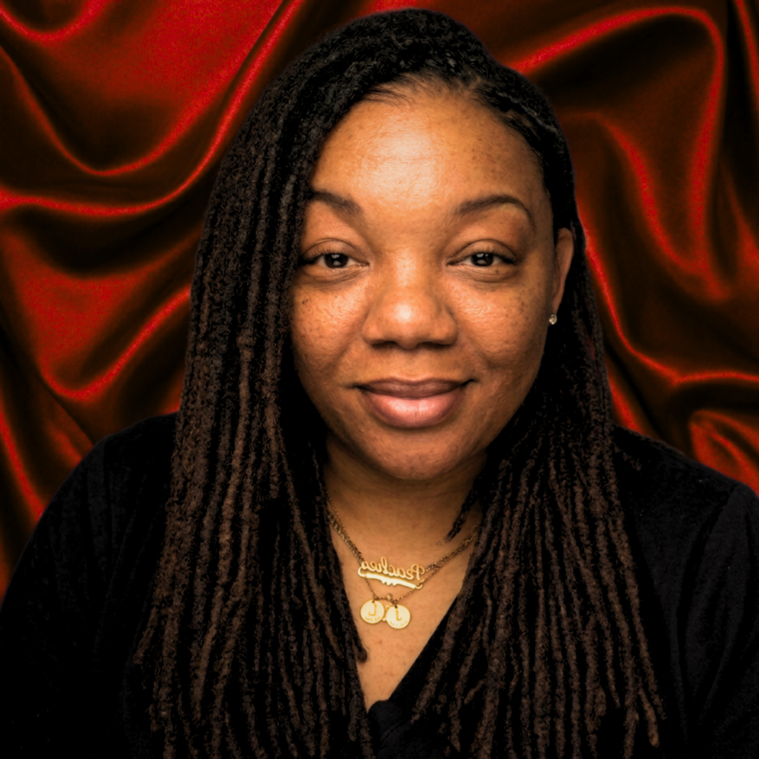 Close-up of a woman's face with braided hair, gold jewelry including necklaces with pendant charms, against a red satin background.