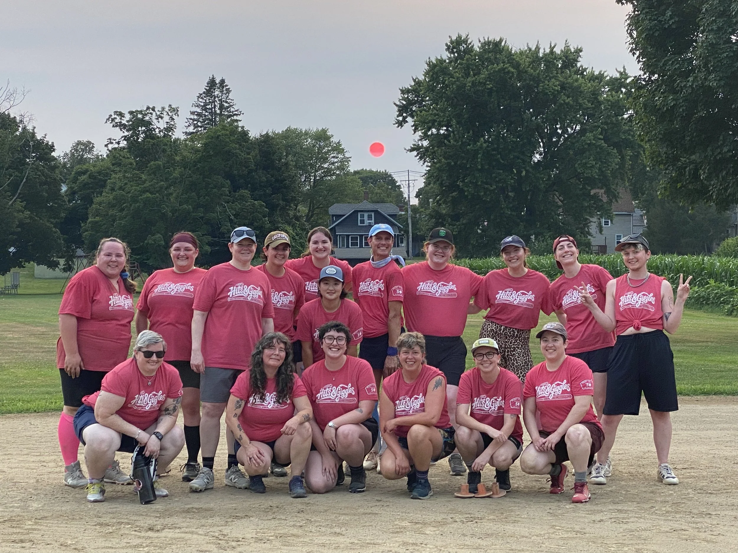 A group of smiling women and girls wearing matching red T-shirts with 'Hits & Giggles' written on them, posing outdoors on a dirt field during the daytime. There are trees, houses, and a red sun or moon in the background.