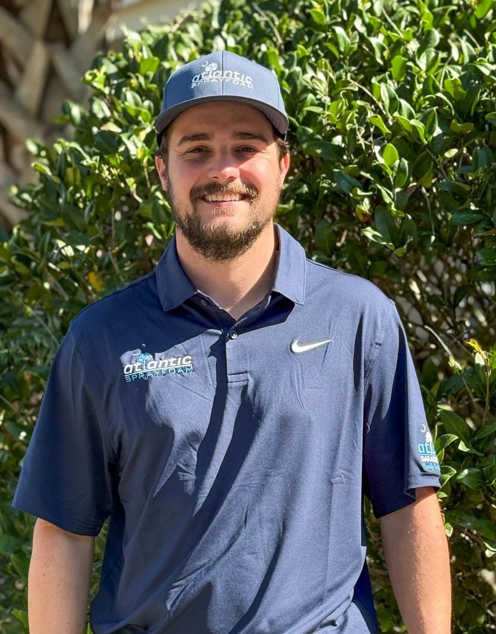 A man smiling outdoors in front of green foliage, wearing a blue cap and a blue polo shirt with logos