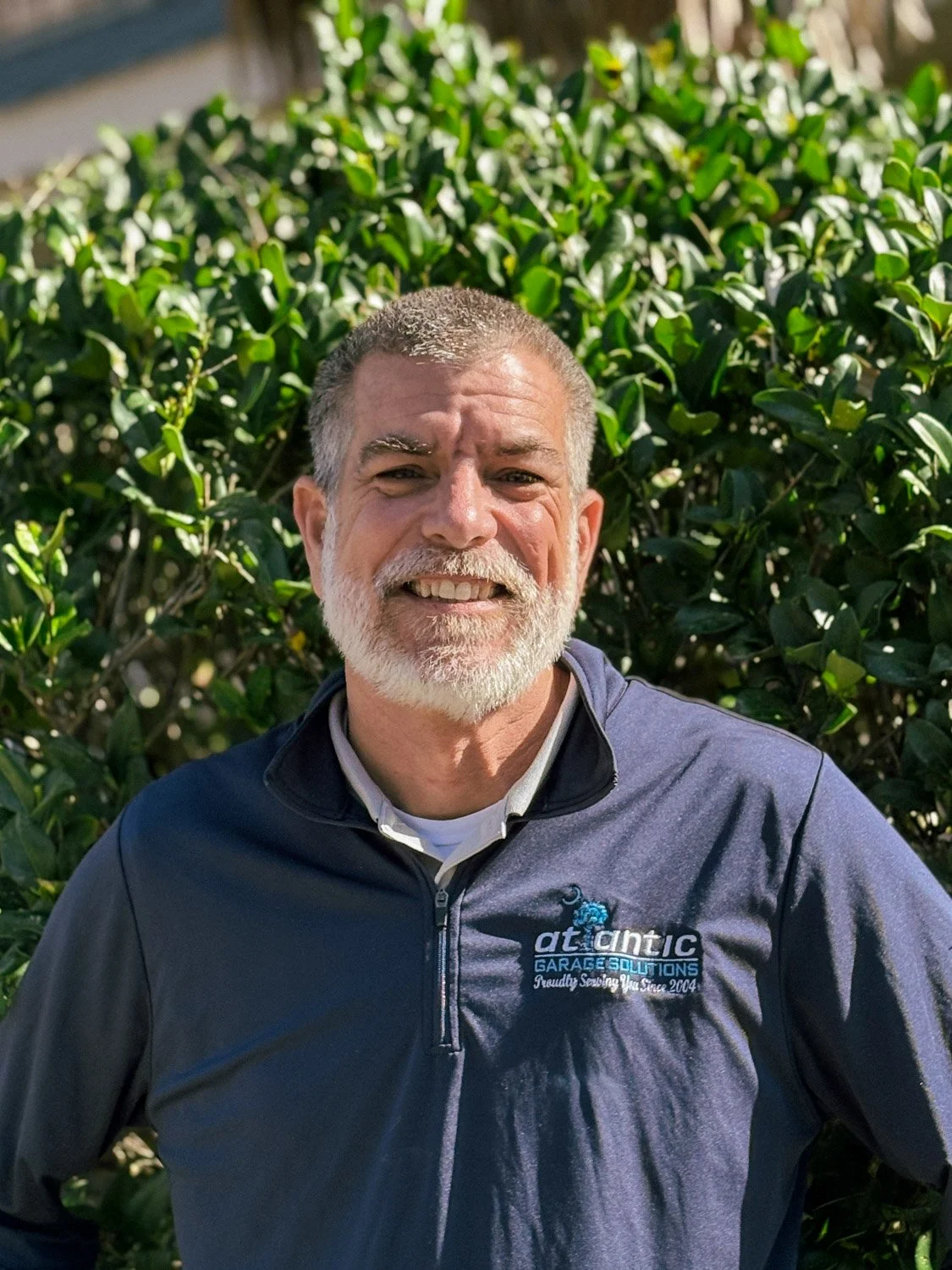 A middle-aged man with gray hair and a beard smiling outdoors in front of green bushes, wearing a dark gray jacket with a logo that reads "Atlantic Garage Solutions" and says "Proudly Serving You Since 2004."
