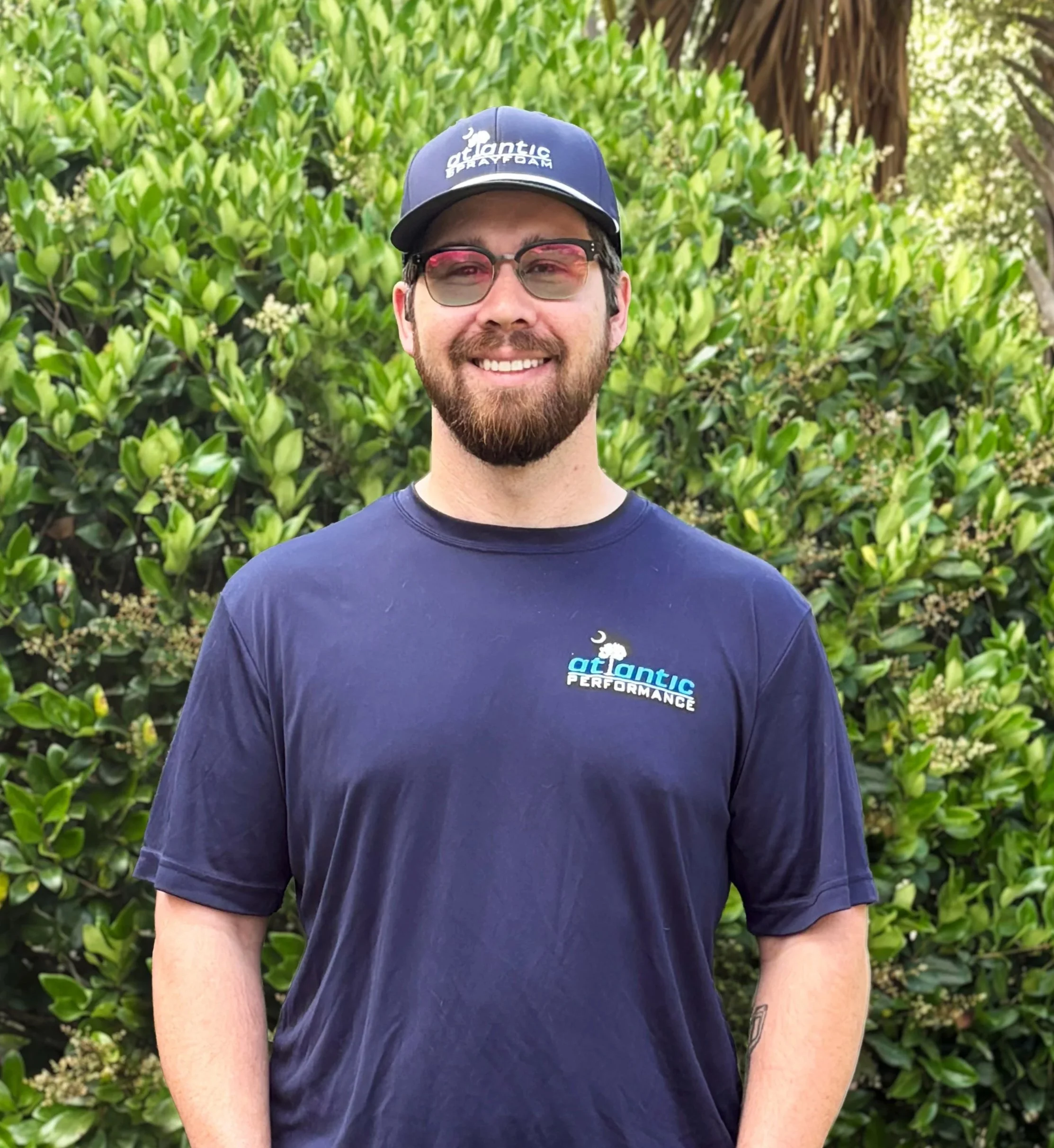 A smiling man with short light brown hair standing outdoors surrounded by blurred green trees, wearing a navy blue polo shirt with sports brand logos.