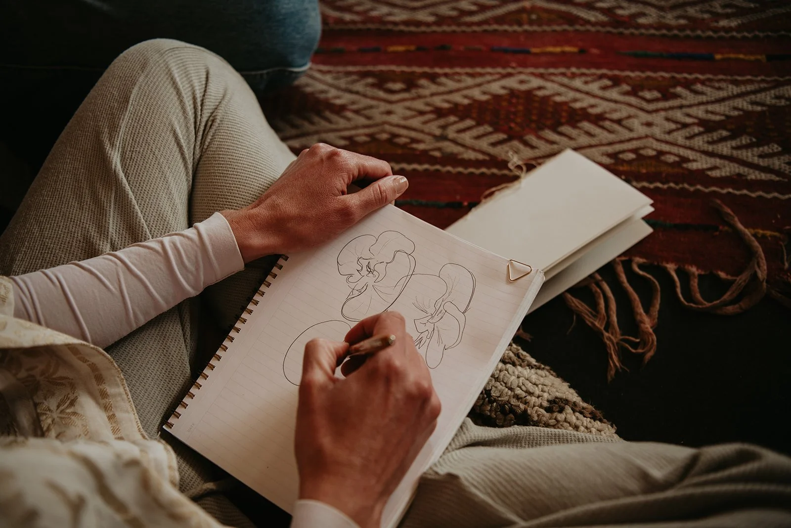 Person drawing abstract face sketches in a notebook, sitting on a patterned rug with legs crossed.