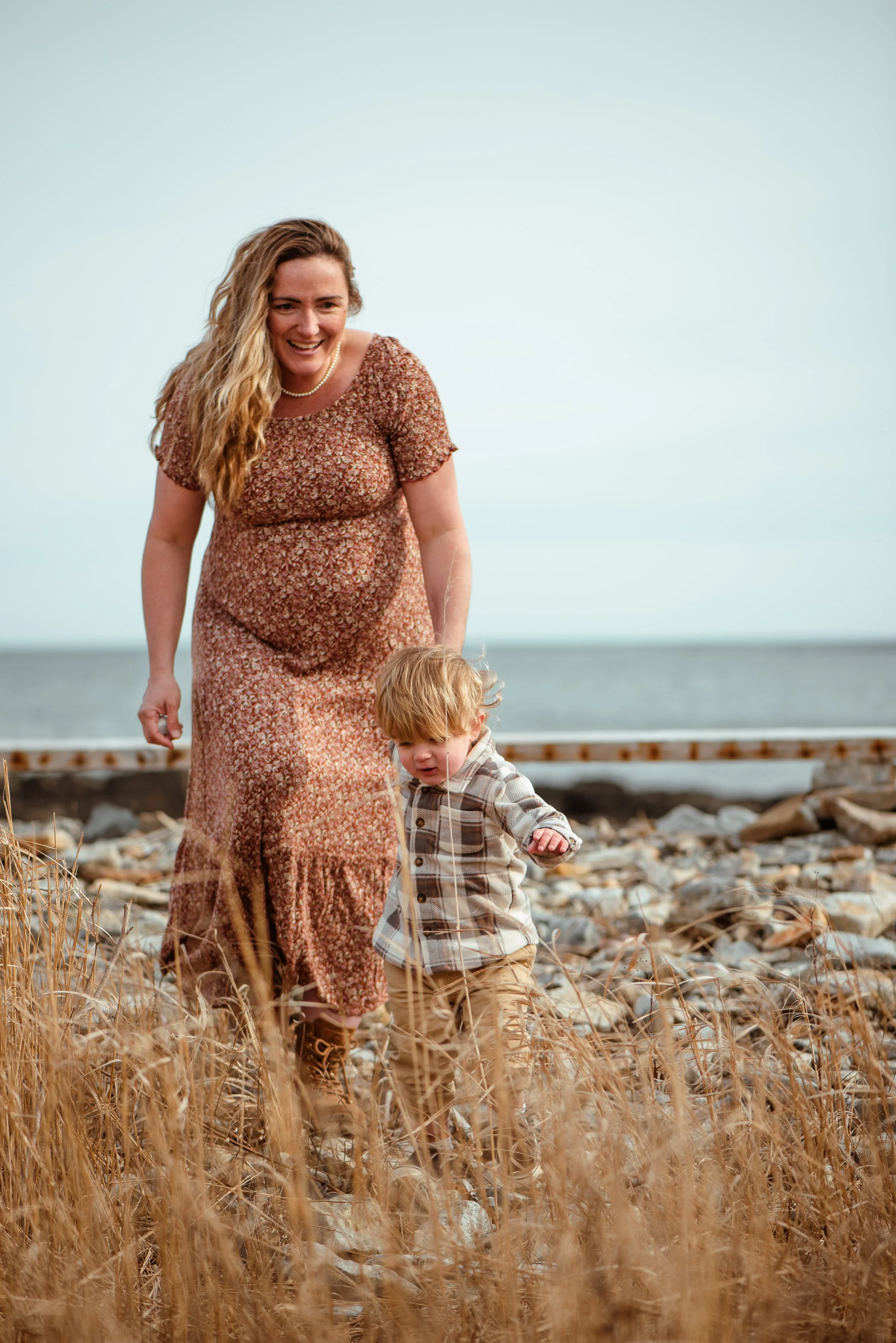 A woman with long blonde hair wearing a floral dress and pearl necklace, smiling as she walks with a young boy in a plaid shirt through a field of tall, dry grasses near a shoreline with a body of water and cloudy sky in the background.