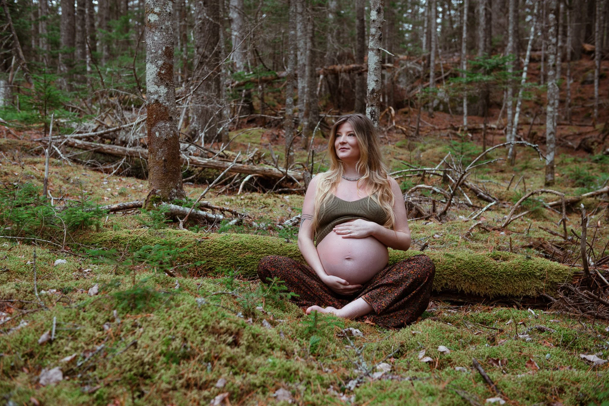 A pregnant woman sitting cross-legged on the forest floor, touching her belly, surrounded by trees and moss.